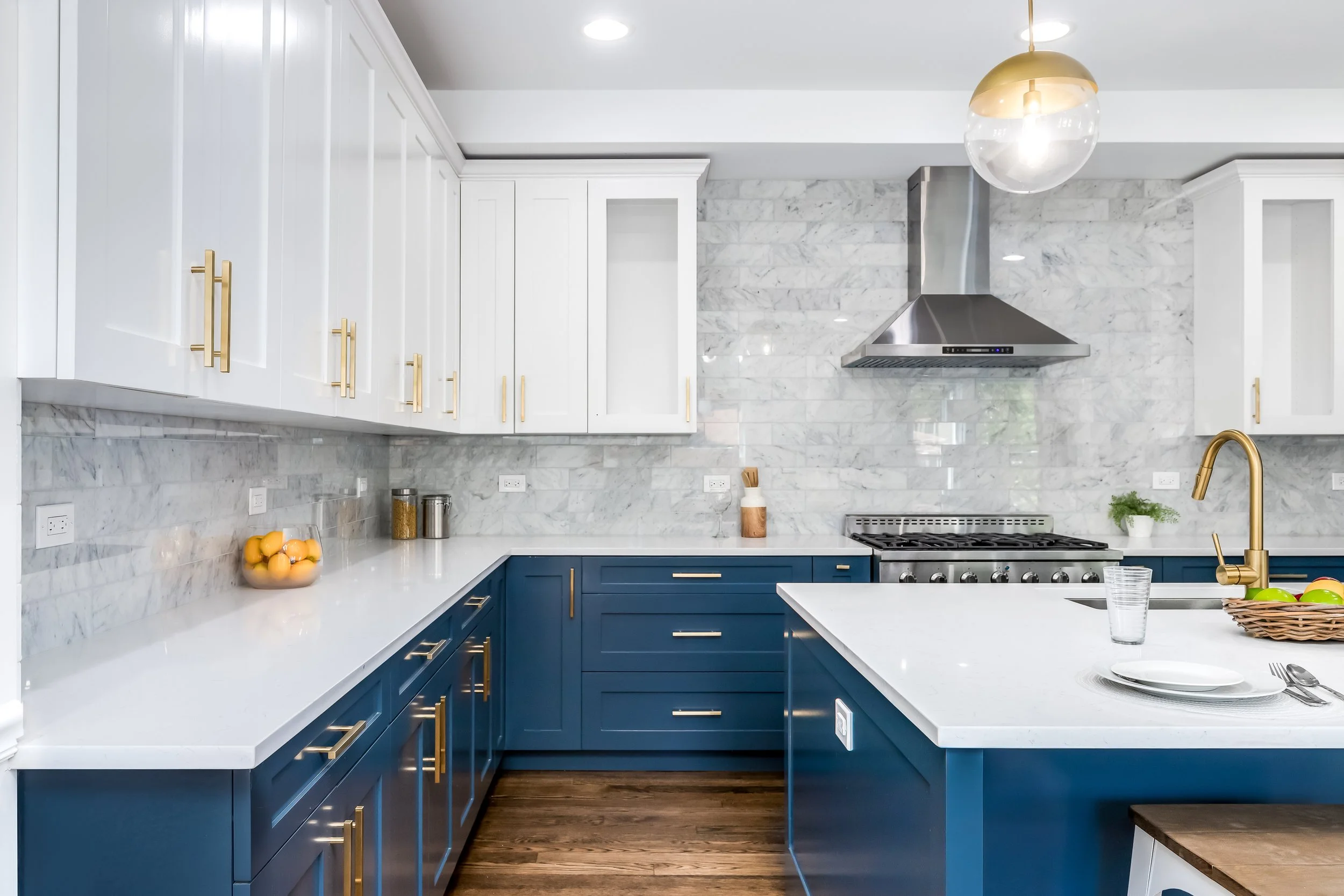 Modern kitchen with white upper cabinets and blue lower cabinets, white countertop, stainless steel range hood, marble backsplash, gold hardware, a basket of green apples, a glass of water, and a gold and glass pendant light.