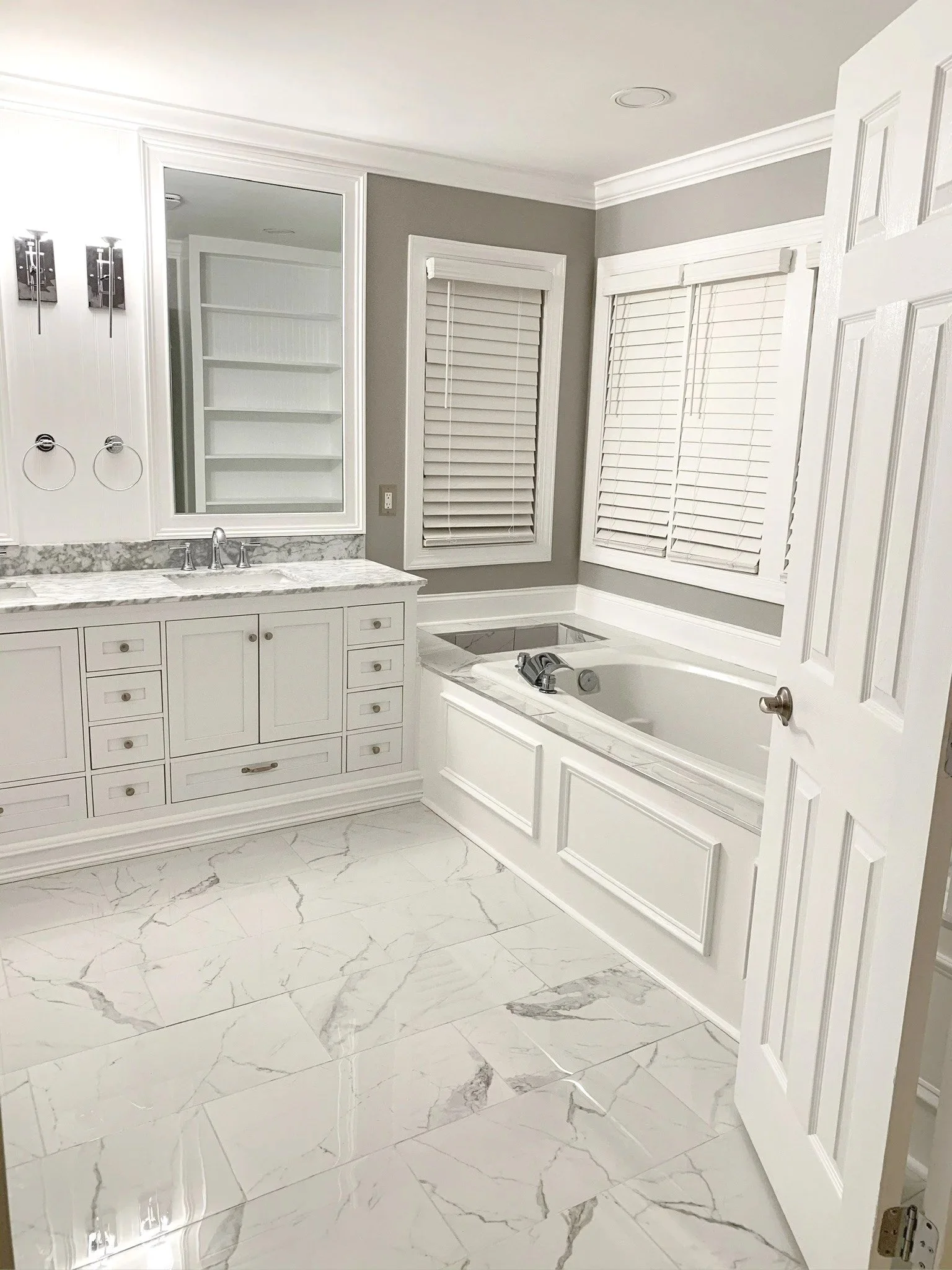 A modern bathroom with white cabinetry, marble countertops, a bathtub, and two windows with white blinds.