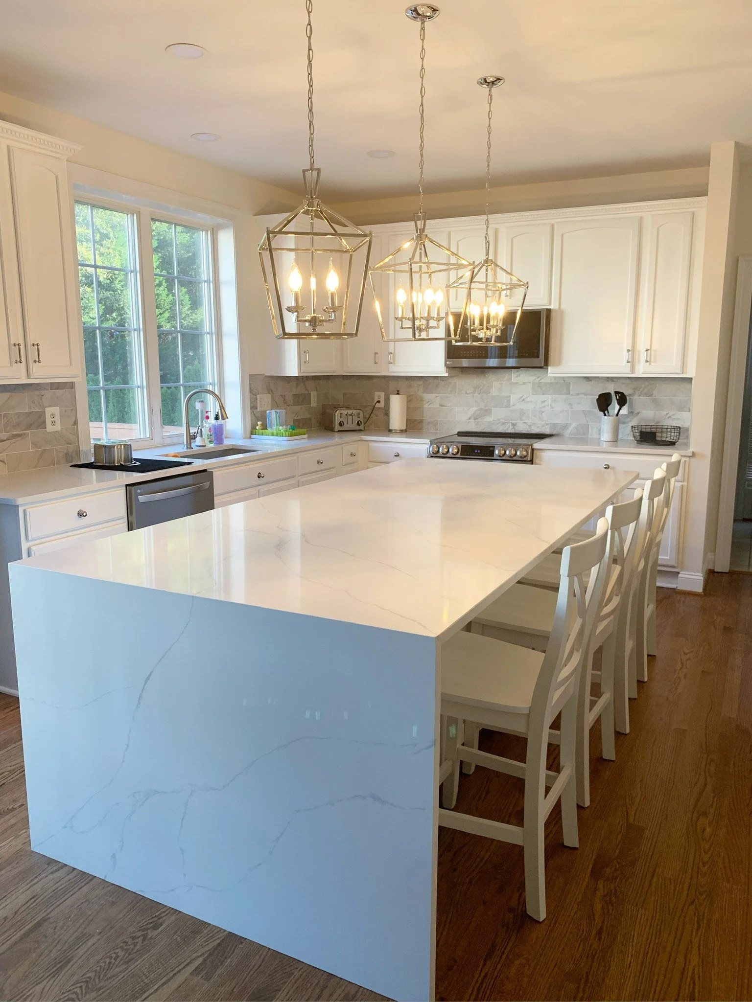 A bright, modern kitchen with white cabinetry, a large white island with a marble-like surface, hardwood floors, and three geometric pendant lights hanging above the island. There is a stainless steel sink beneath a large window, and appliances including a stove, microwave, and toaster. The kitchen has neutral-colored backsplash tiles.