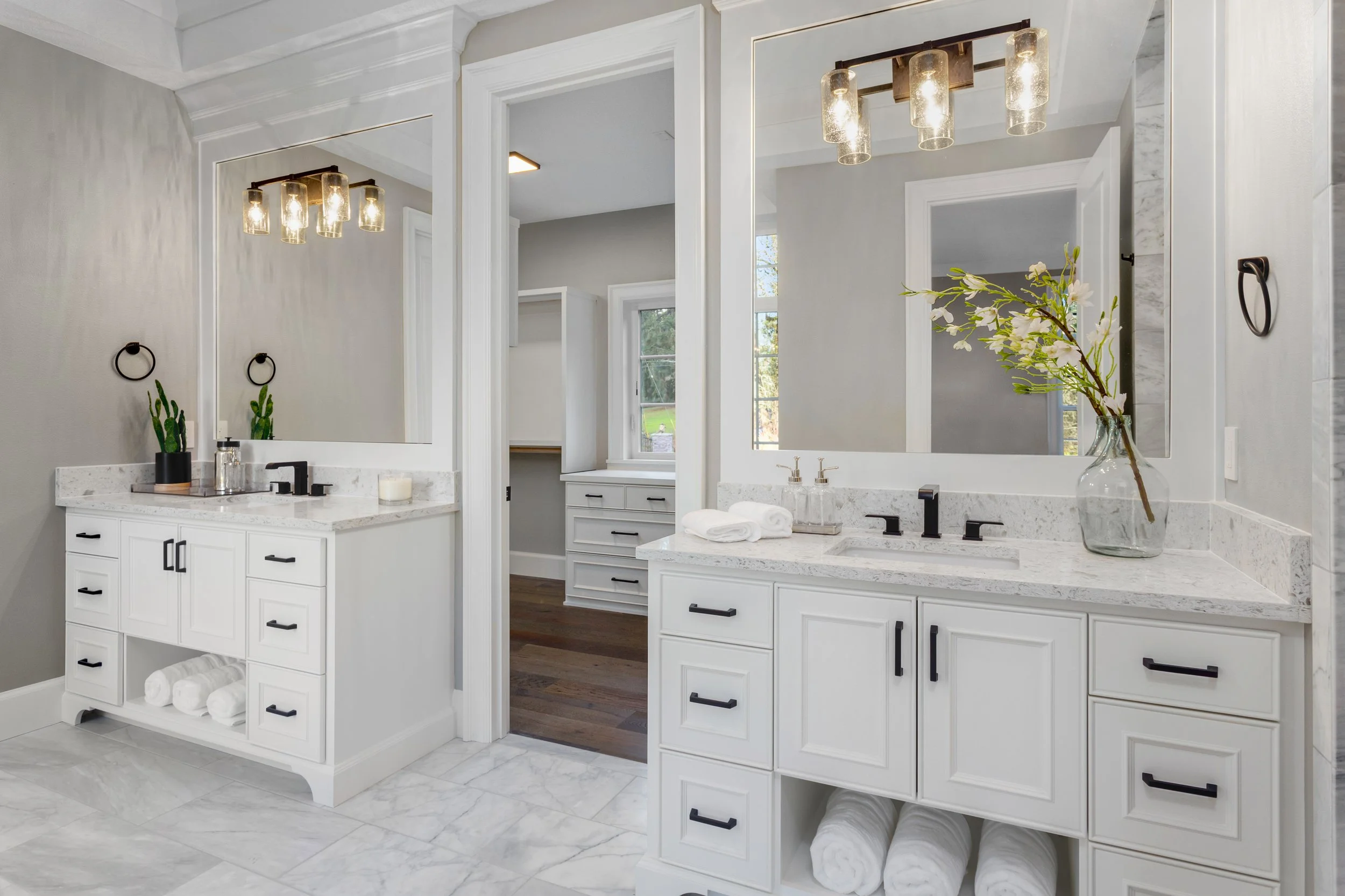 Modern bathroom with dual white vanities, large mirrors, black fixtures, and decorative items including green plants and flowers, illuminated by contemporary light fixtures.