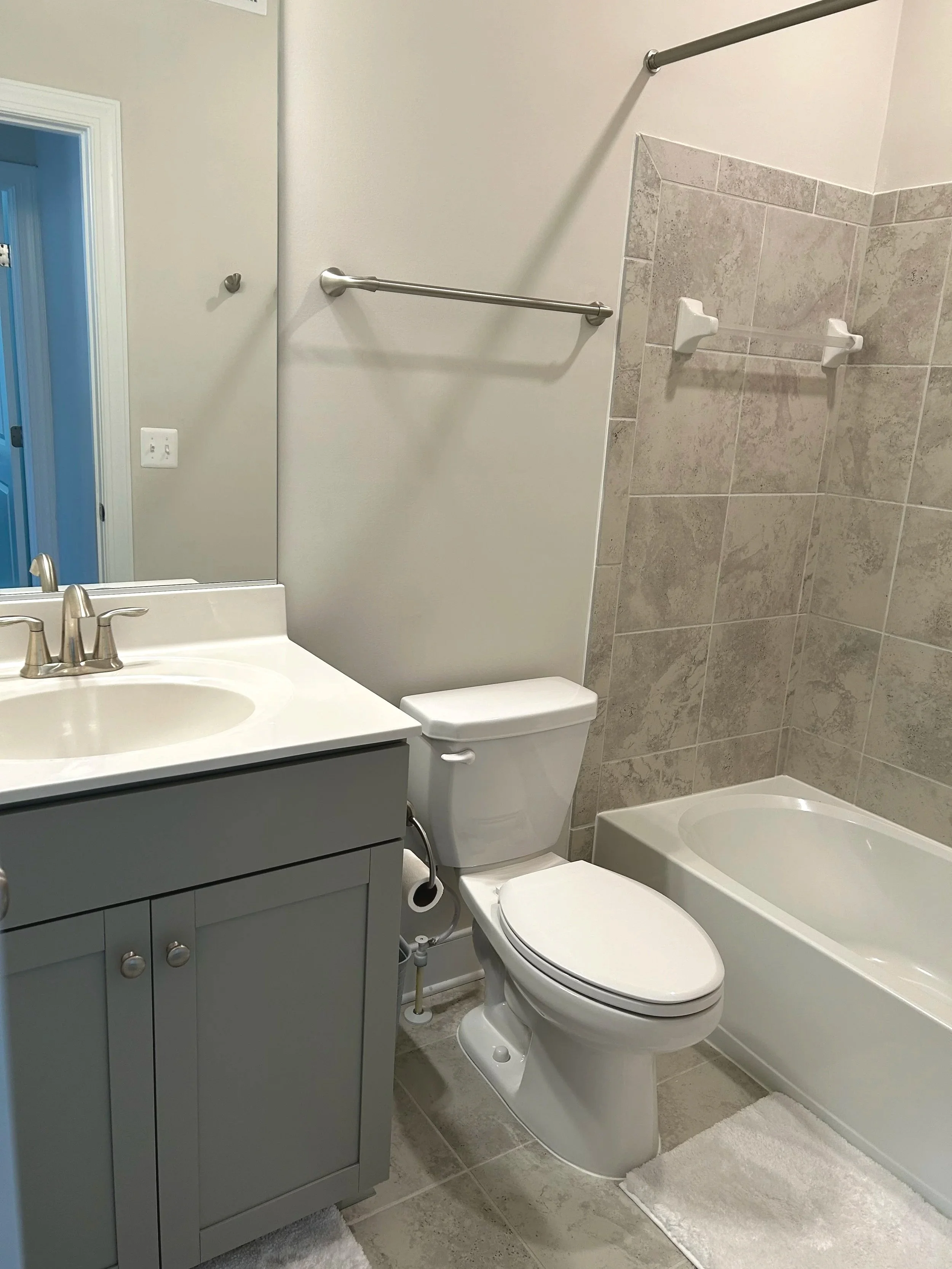 A small bathroom with a vanity, toilet, and bathtub. The vanity has a gray cabinet with a sink and faucet. The toilet is positioned next to the vanity, and the bathtub is along the wall with two white soap dishes. There are two towel bars on the wall, a large mirror above the sink, and a doorway with blue trim visible in the background.