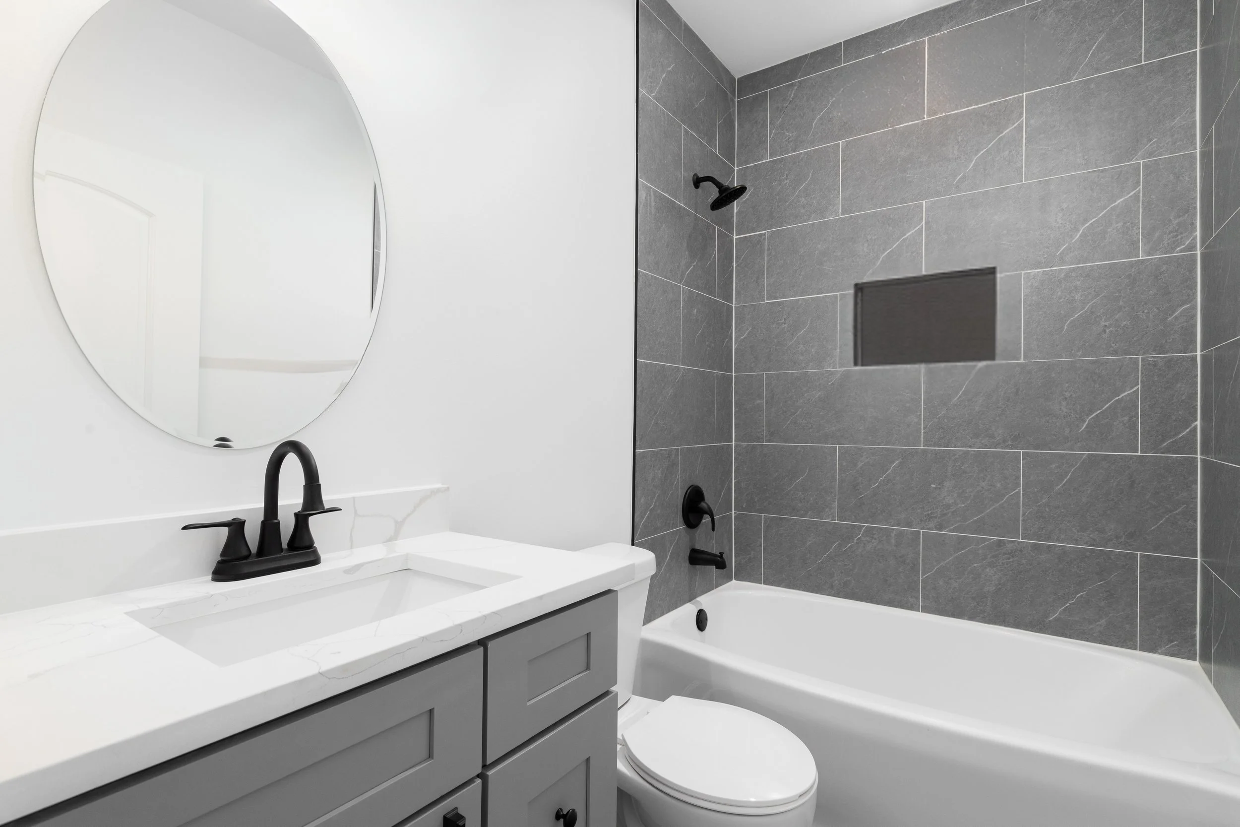 Modern bathroom with white vanity, black faucet, oval mirror, gray tiled wall in shower, white bathtub, and white toilet.