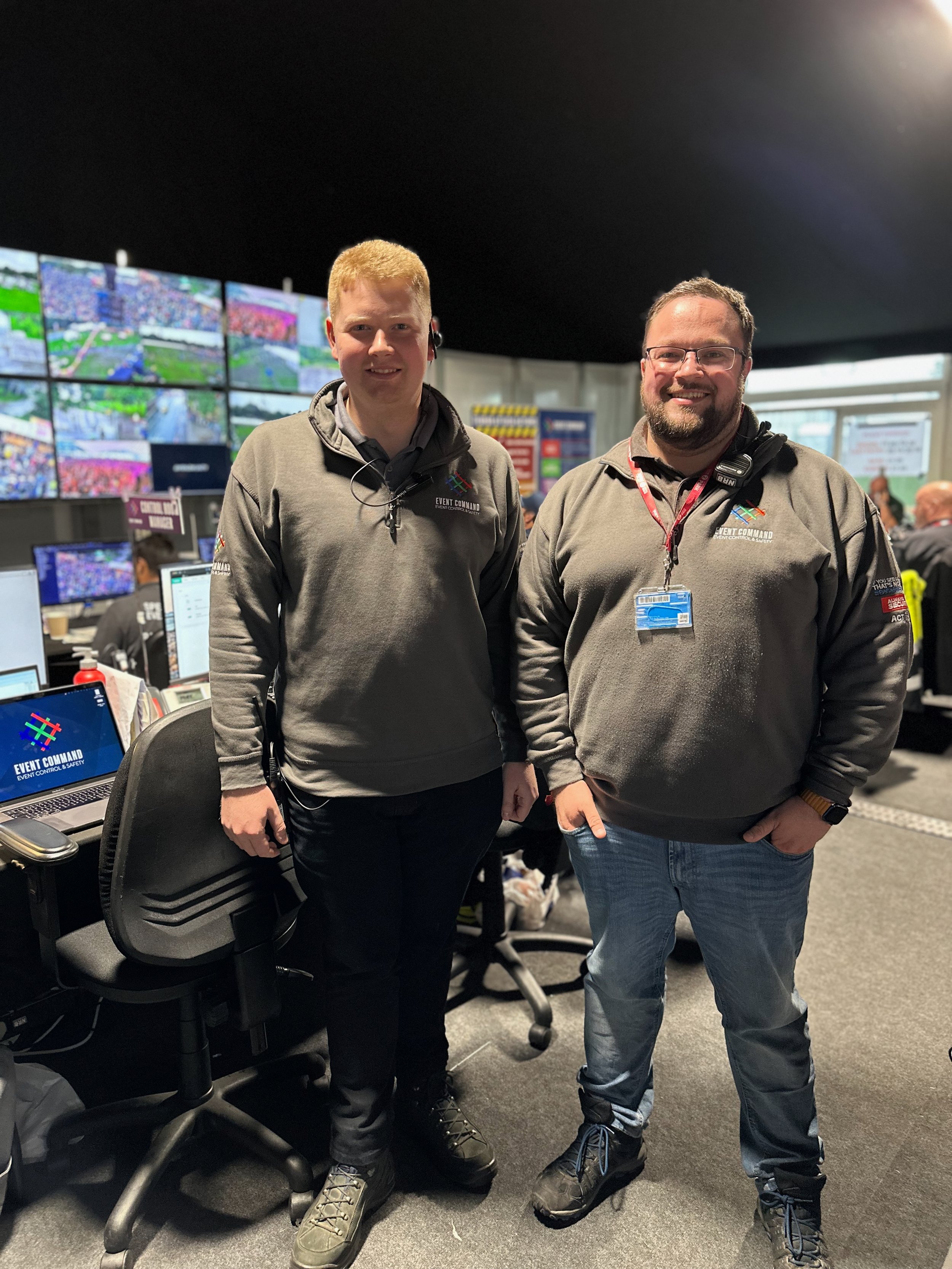 Two Event Command team members stand in an event control room wearing branded grey fleeces, with CCTV monitor walls, computer workstations and radio communications equipment visible behind them.