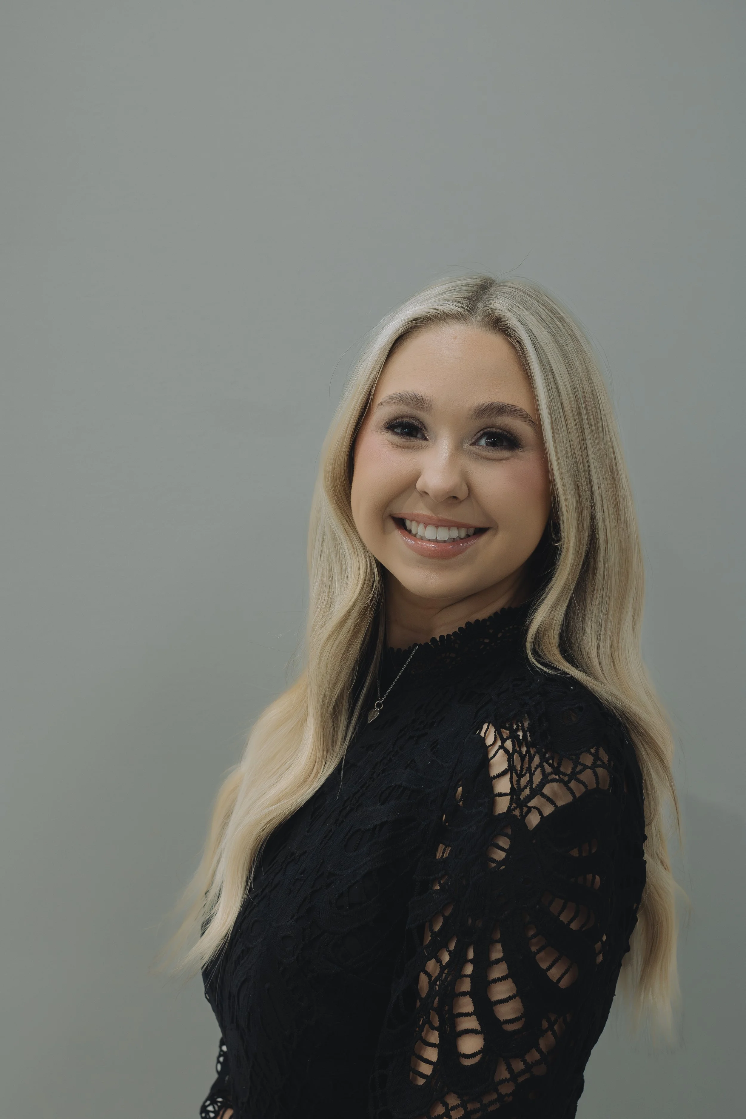 A young woman with long blonde hair smiling at the camera, wearing a black lace top, standing against a plain gray background.