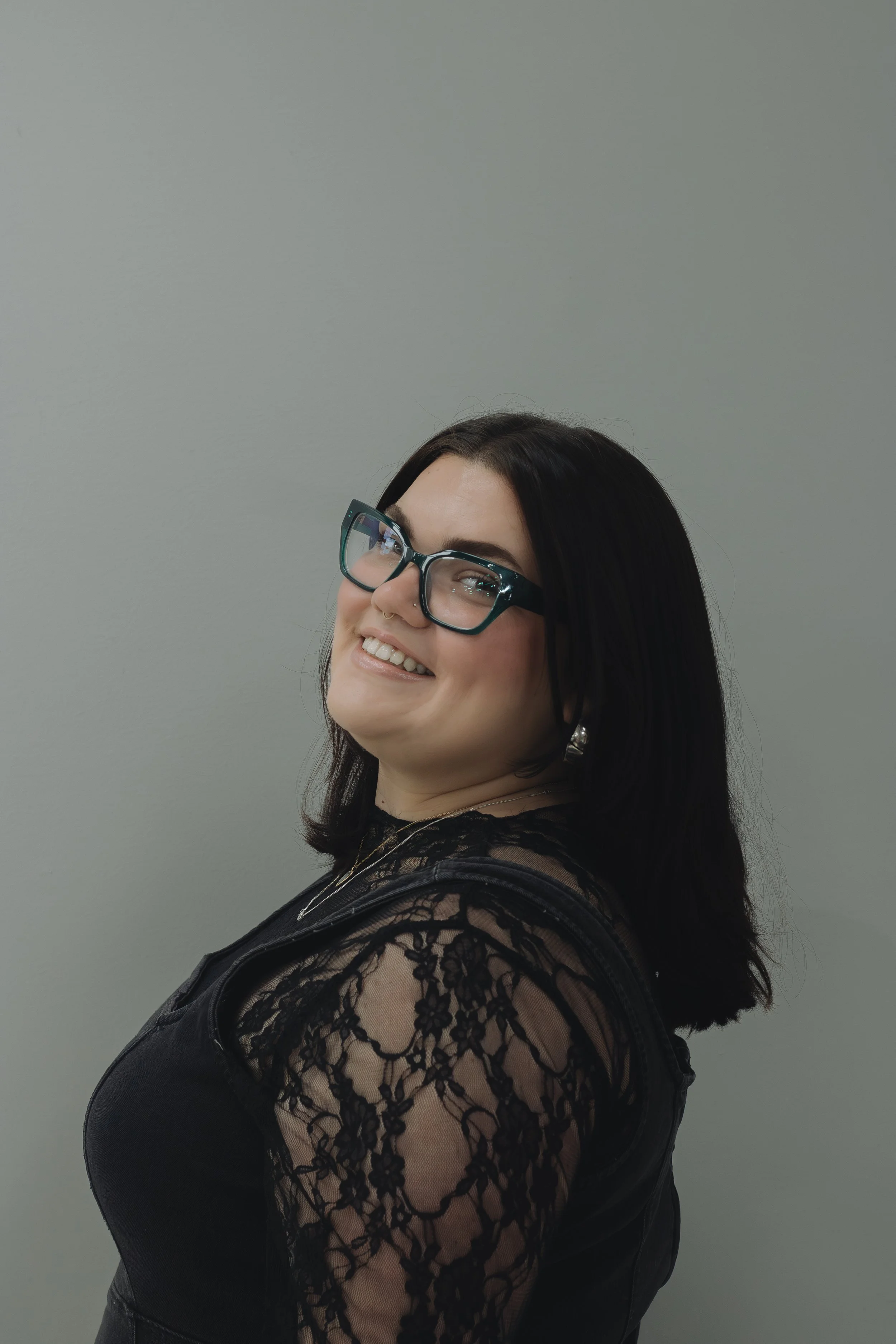 Smiling woman with dark hair and glasses, wearing a black lace top, against a plain gray background.