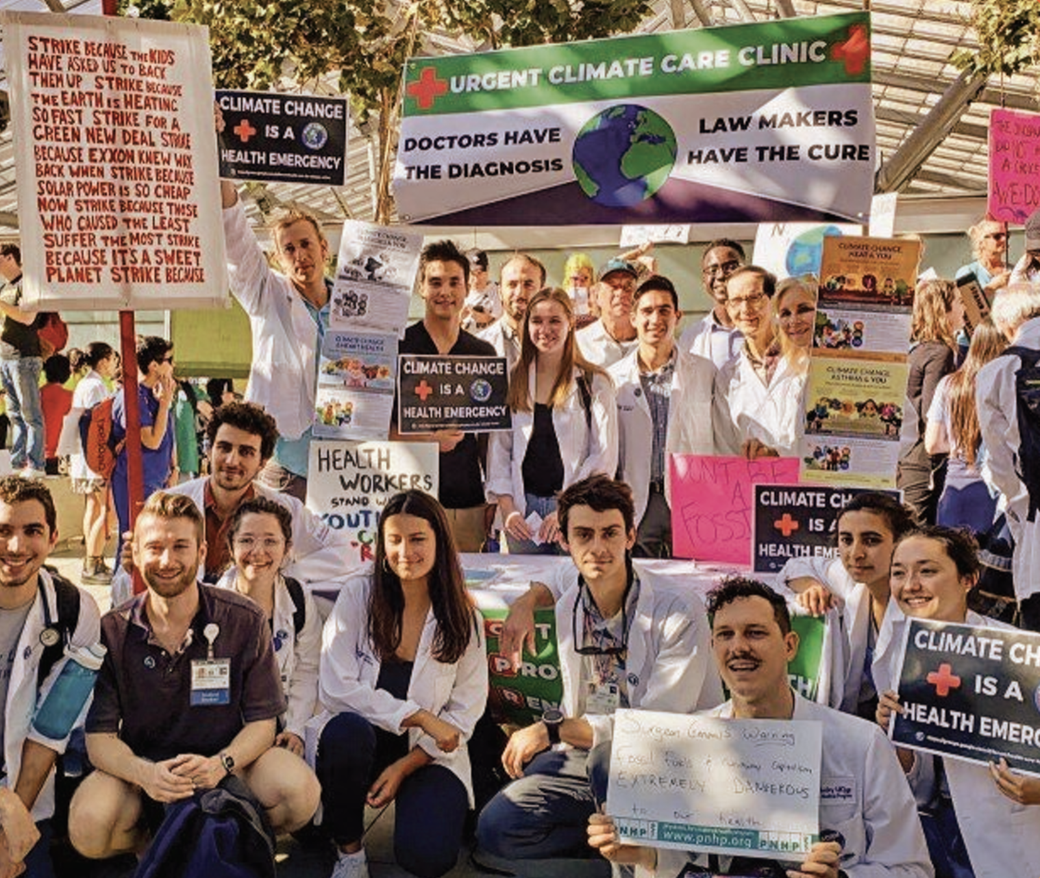 Group of people at a climate and healthcare protest holding signs and banners, including one reading 'Urgent Climate Care Clinic' and another about health emergencies and climate change.