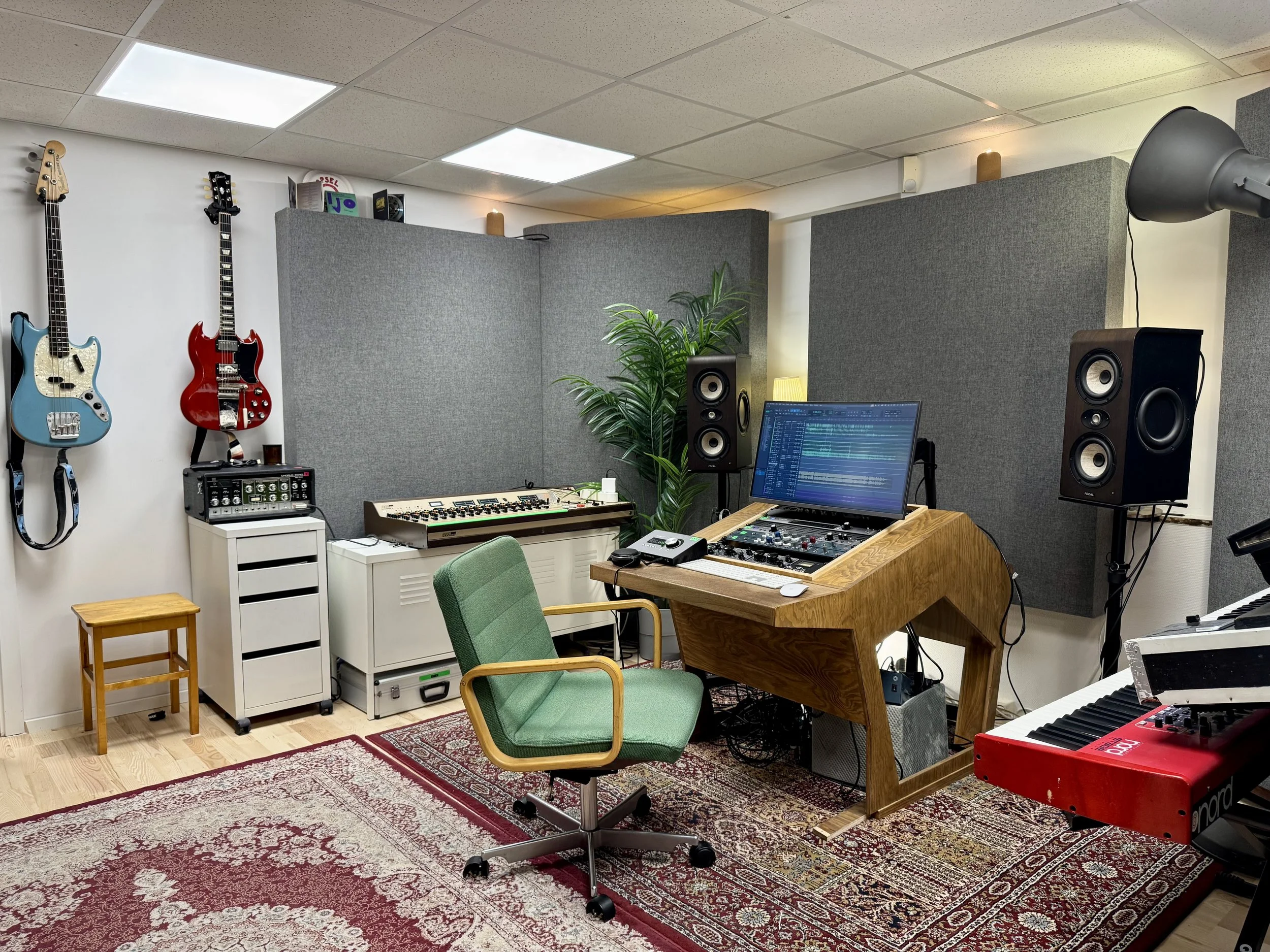 Innspillingsstudio med musikkutstyr, inkludert gitarer på veggen, høyttalere, keyboard, datamaskin, studiobord og stol, med akustiske paneler på veggene