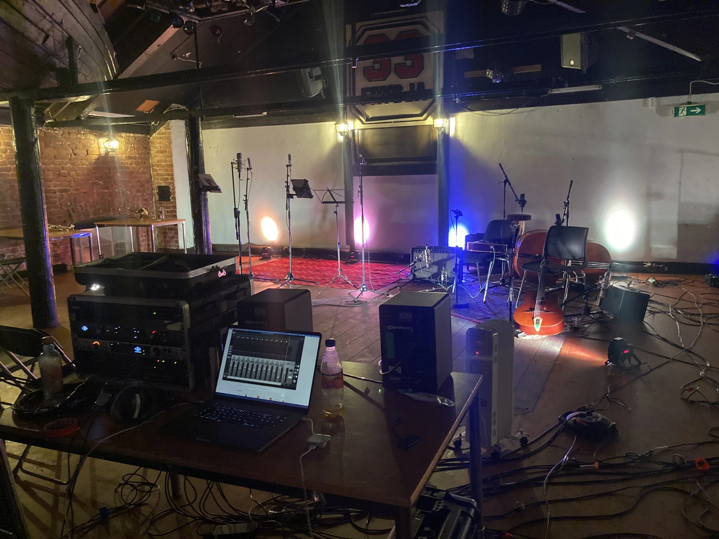 En scene for live musikk med mikrofoner, gitarer, og musikkinstrumenter, opplyst med fargede lys. Bak scenen er det en bruttvegg og en stor skjerm. Frontruten er utstyrt med lyd- og lysutstyr, kabler og en datamaskin.