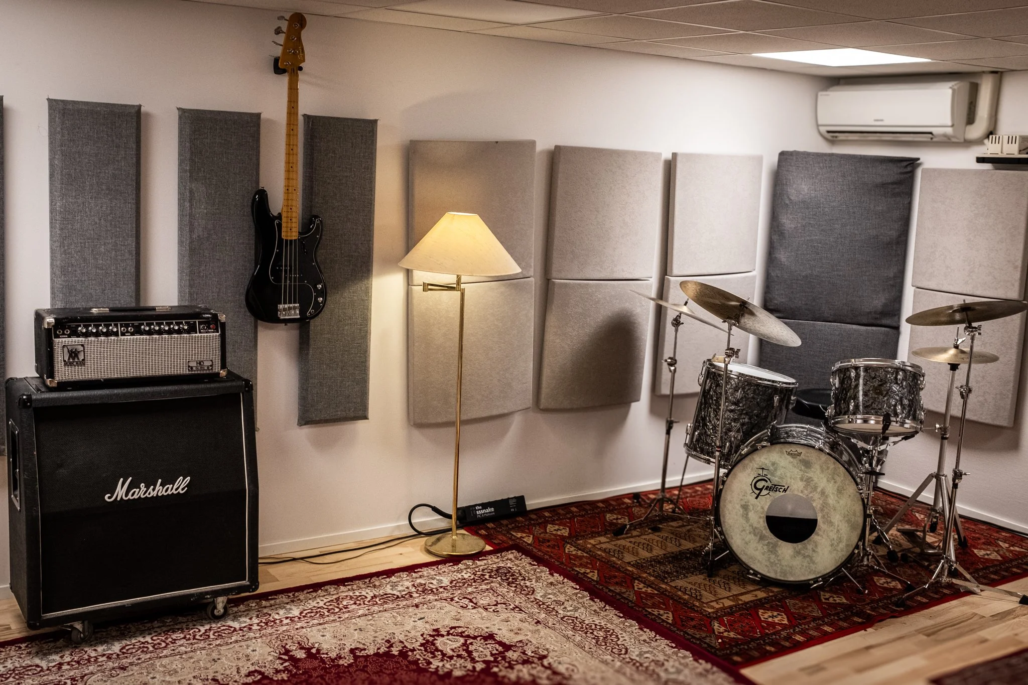 Musikklokale med gitar, forsterker, trommesett, veggplater, gulvlampe, og lydutstyr.