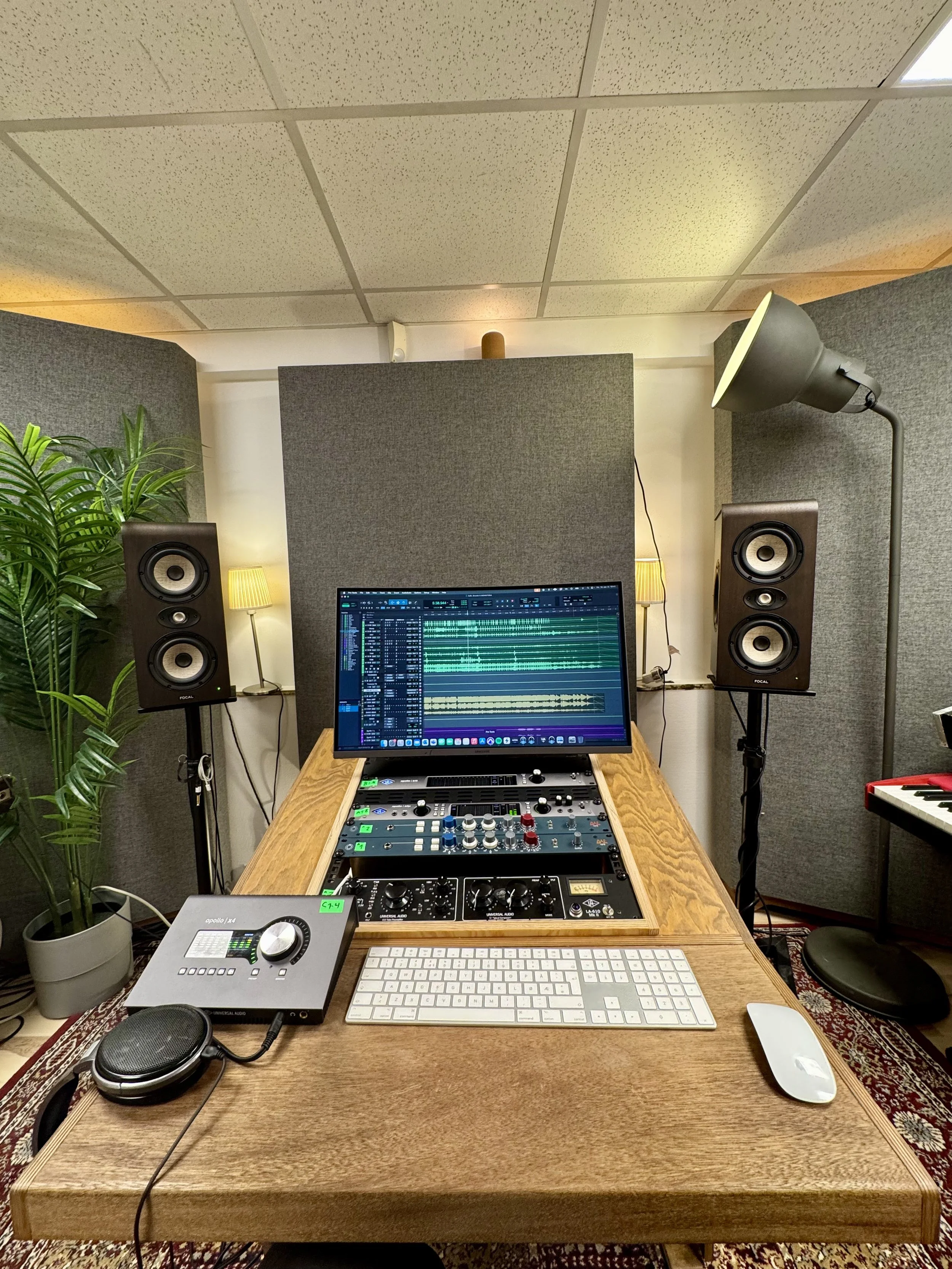 Innspillingsrom med dataskjerm som viser digitalt musikkproduksjonsprogram, studiohøyttalere, MIDI-tastatur, koppholder, hodetelefoner, og lyspærer, med grønne planter og lydplater på veggen.