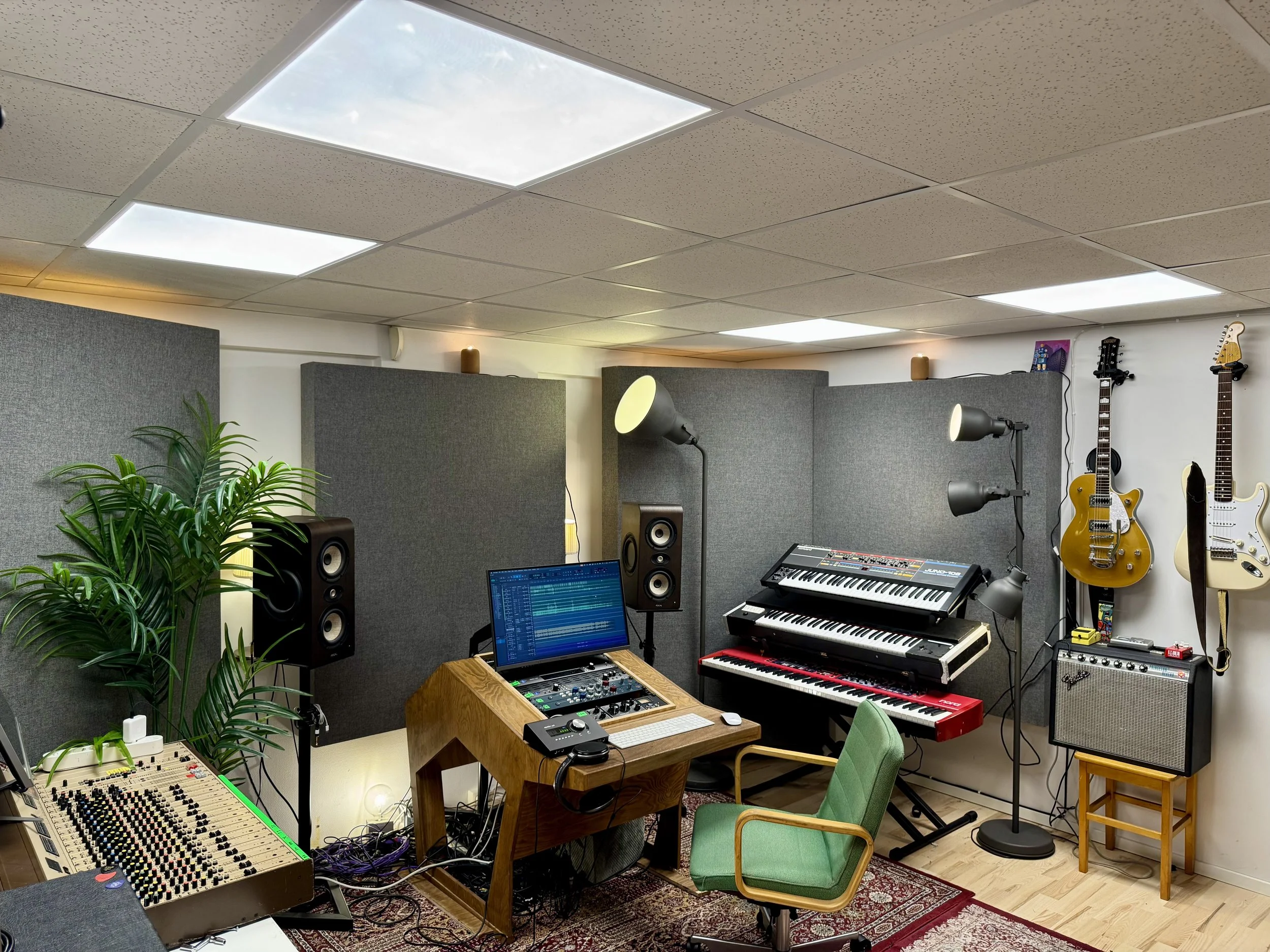 Innendørs musikkstudio med gitarer på veggen, keyboards, monitorer, miksere, stoler og en stor plante.