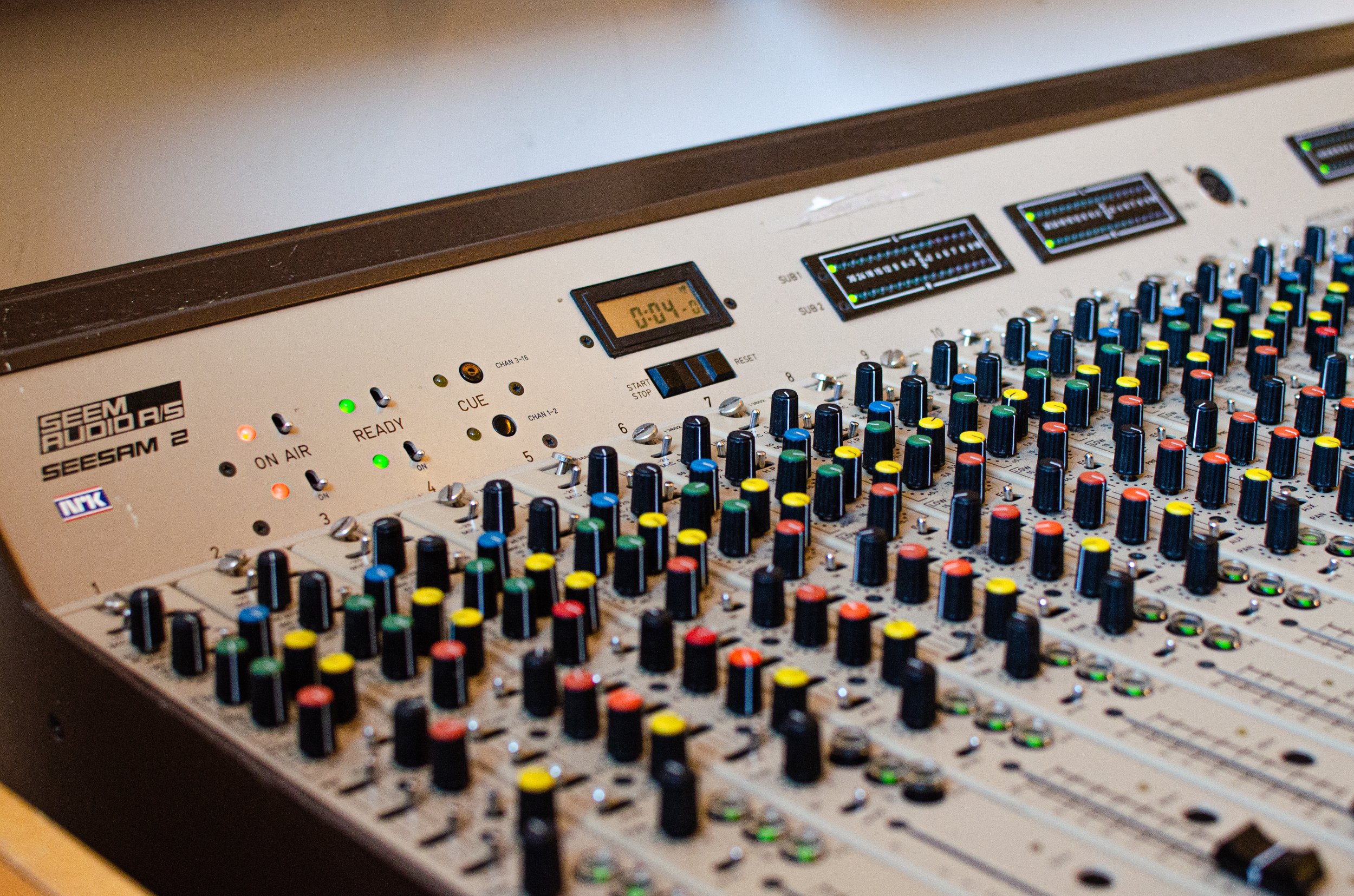 Lyse beige lydmikser med mange fargede knapper og spaker, samt små skjermer som viser signalnivåer og tidsur.