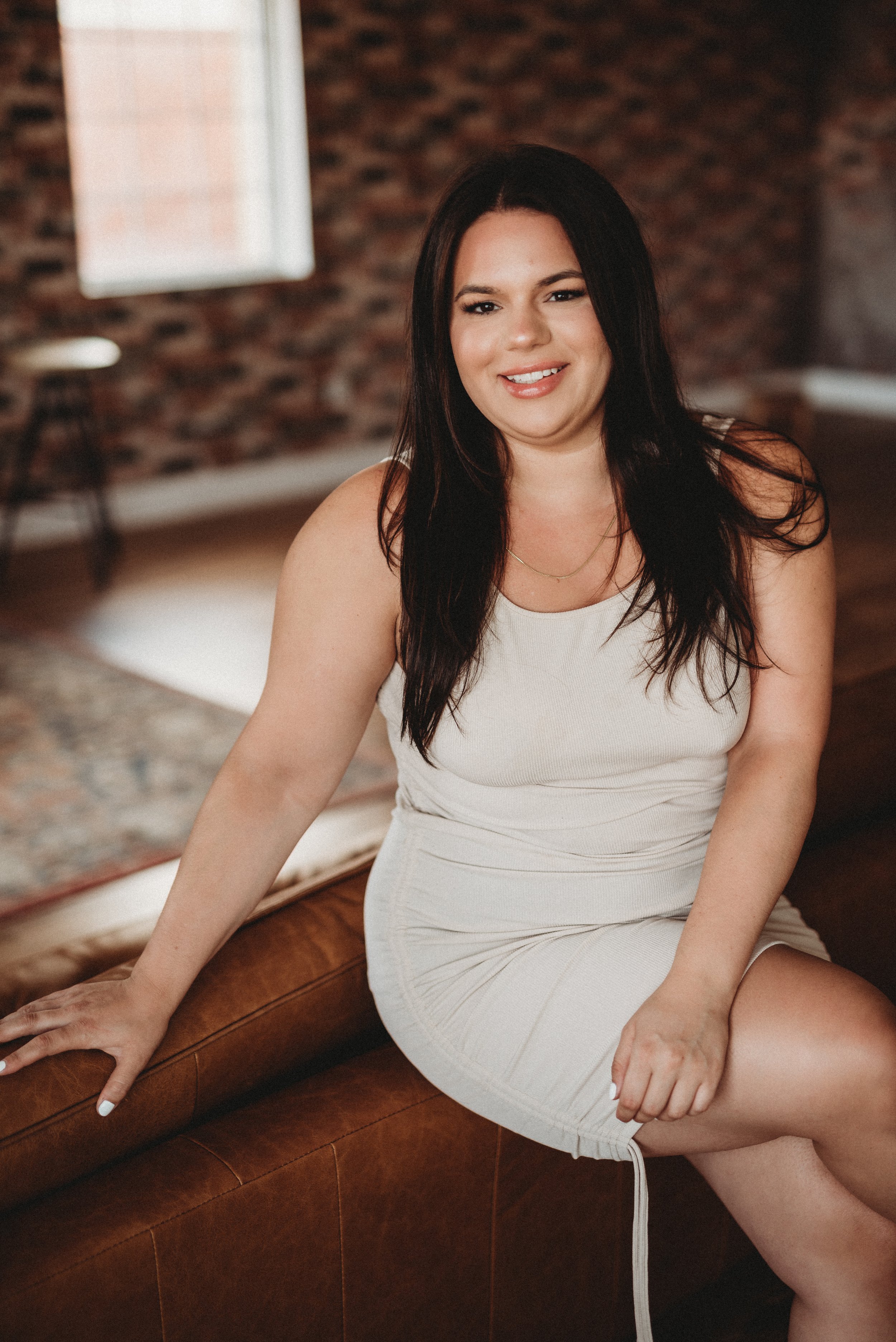 Woman in a light beige dress sitting on a brown couch in a room with brick walls and a window.