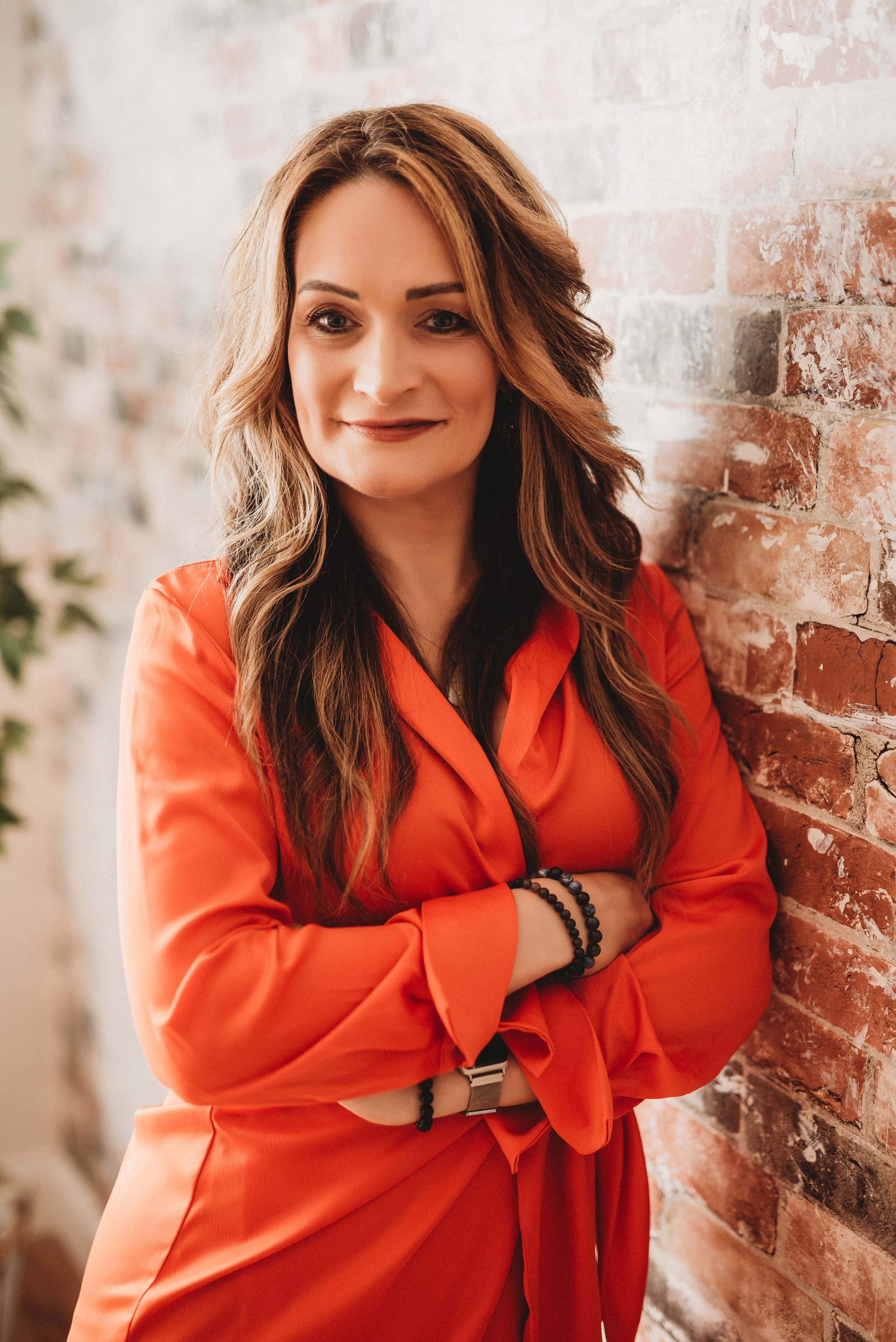 Woman in orange blouse leaning against a brick wall, smiling with arms crossed.