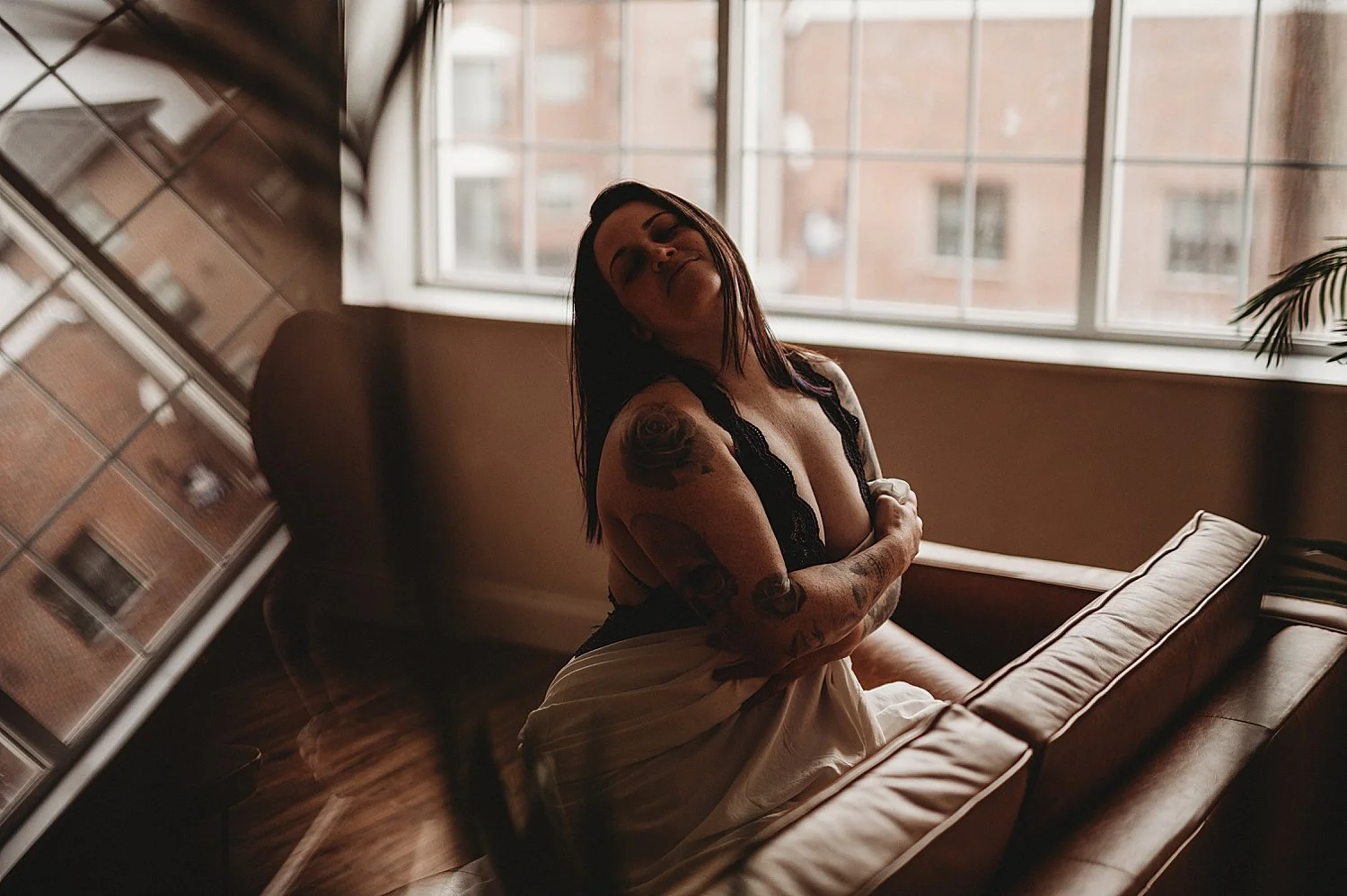  brunette kneels on leather couch in bodysuit by Hannah Mac Studios 