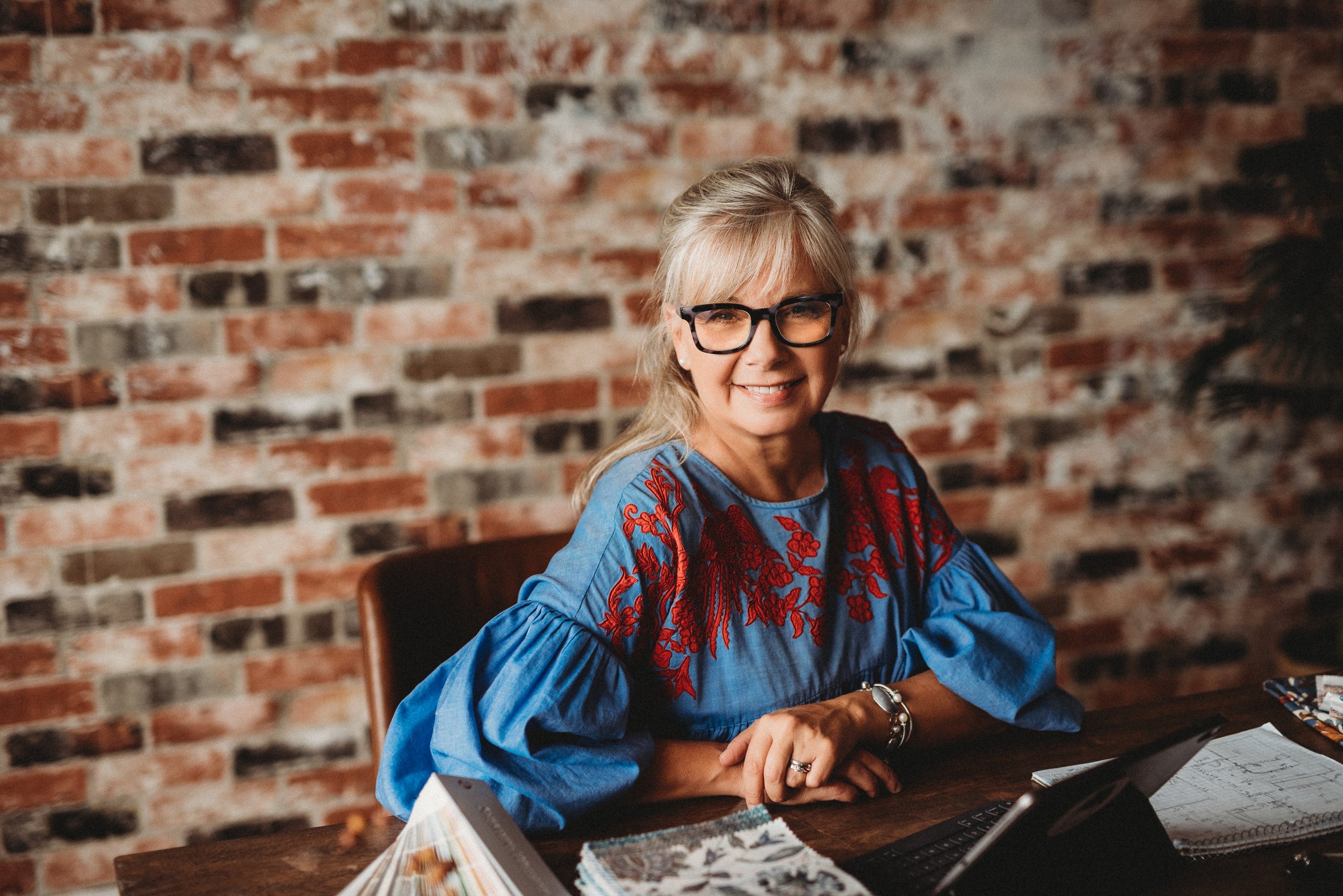 Woman with glasses and long hair sitting at a desk with brick wall background.
