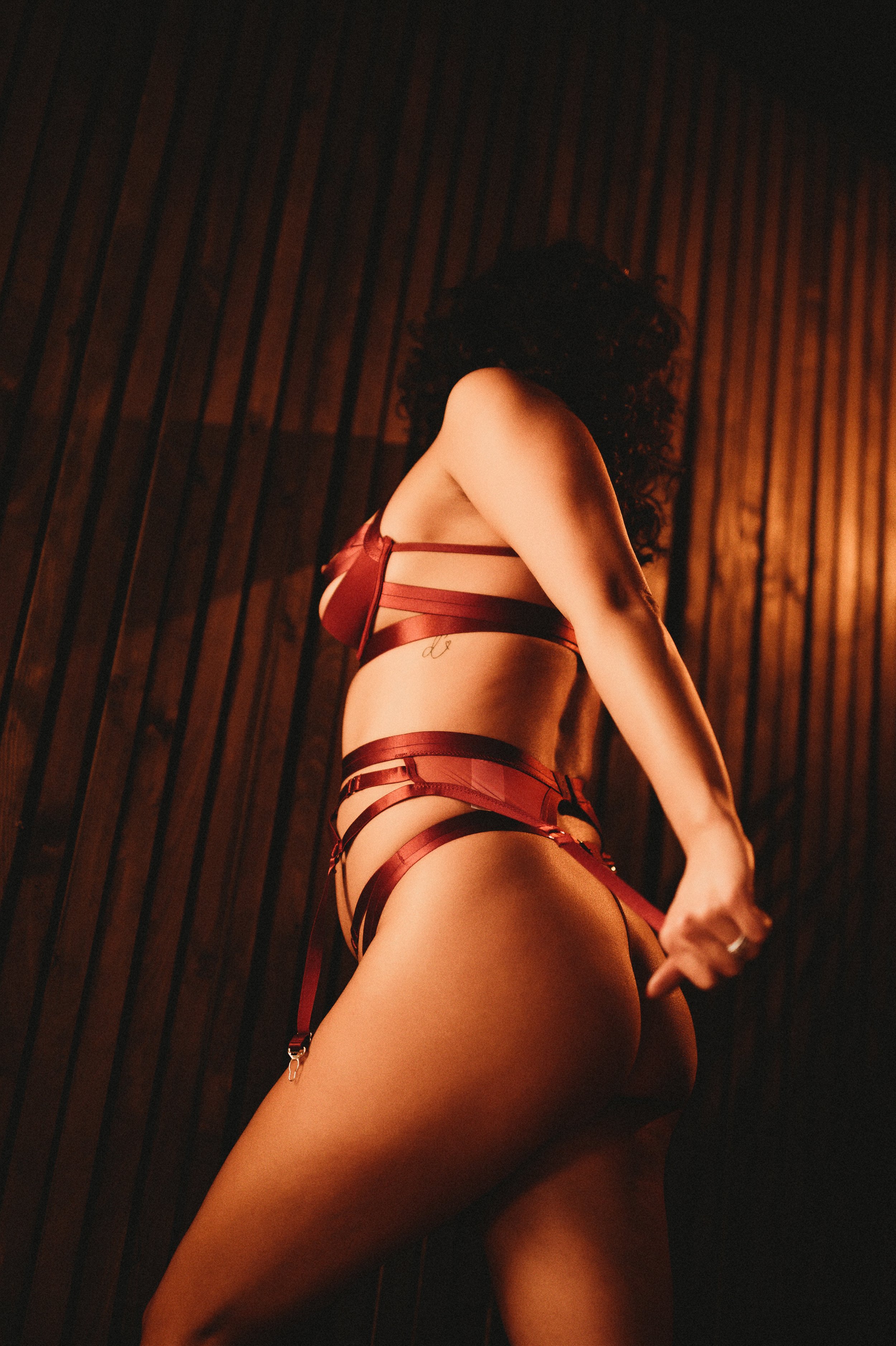 Person wearing strappy red lingerie in dim, warm lighting against a wooden background.