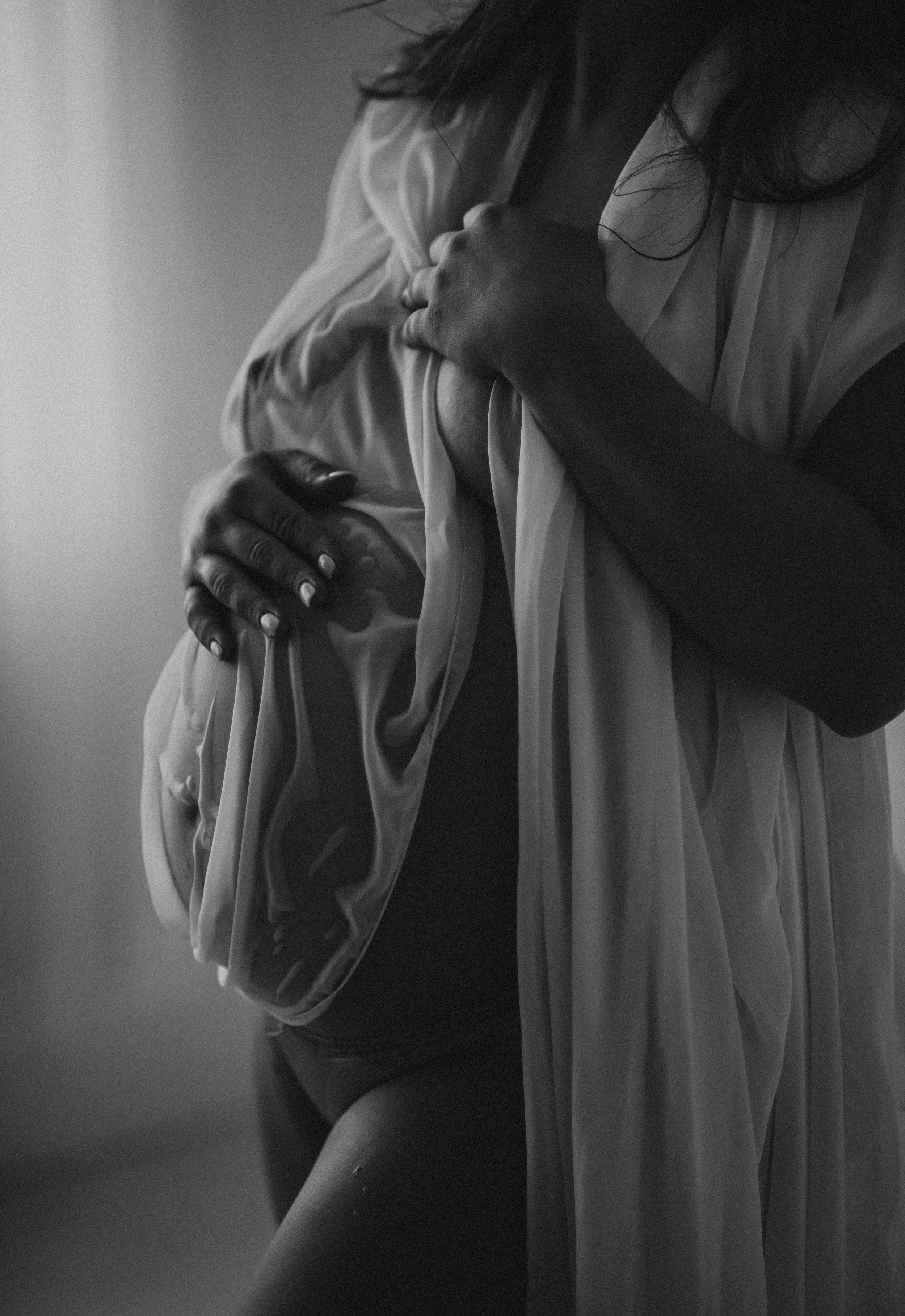Black and white photo of a pregnant woman holding her belly and wearing a sheer fabric draped over her shoulder.