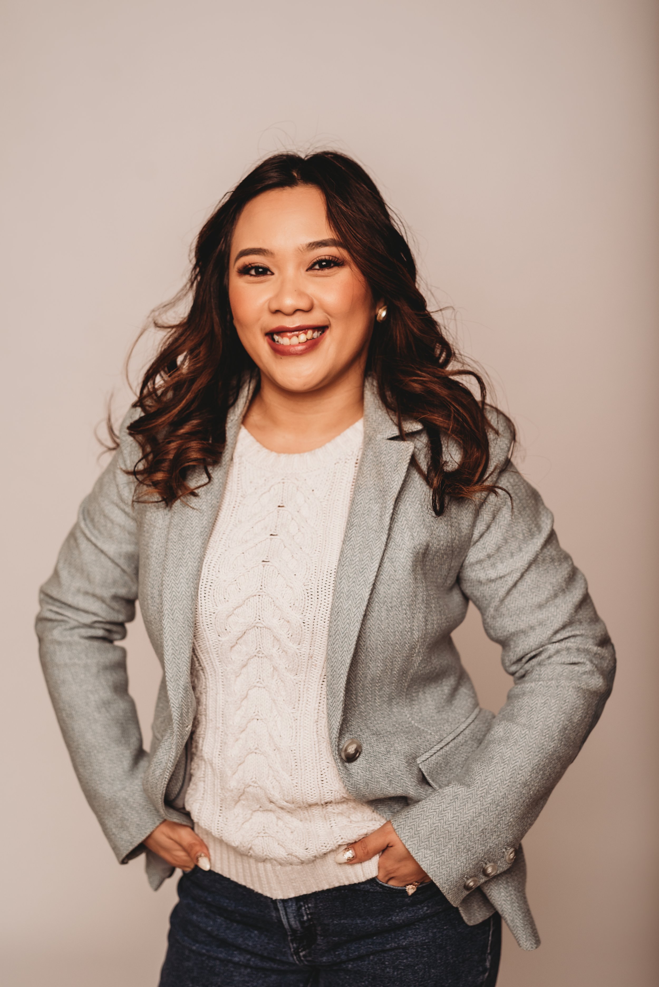 Smiling person wearing a gray blazer and white sweater with hands on hips, posed against a neutral background.