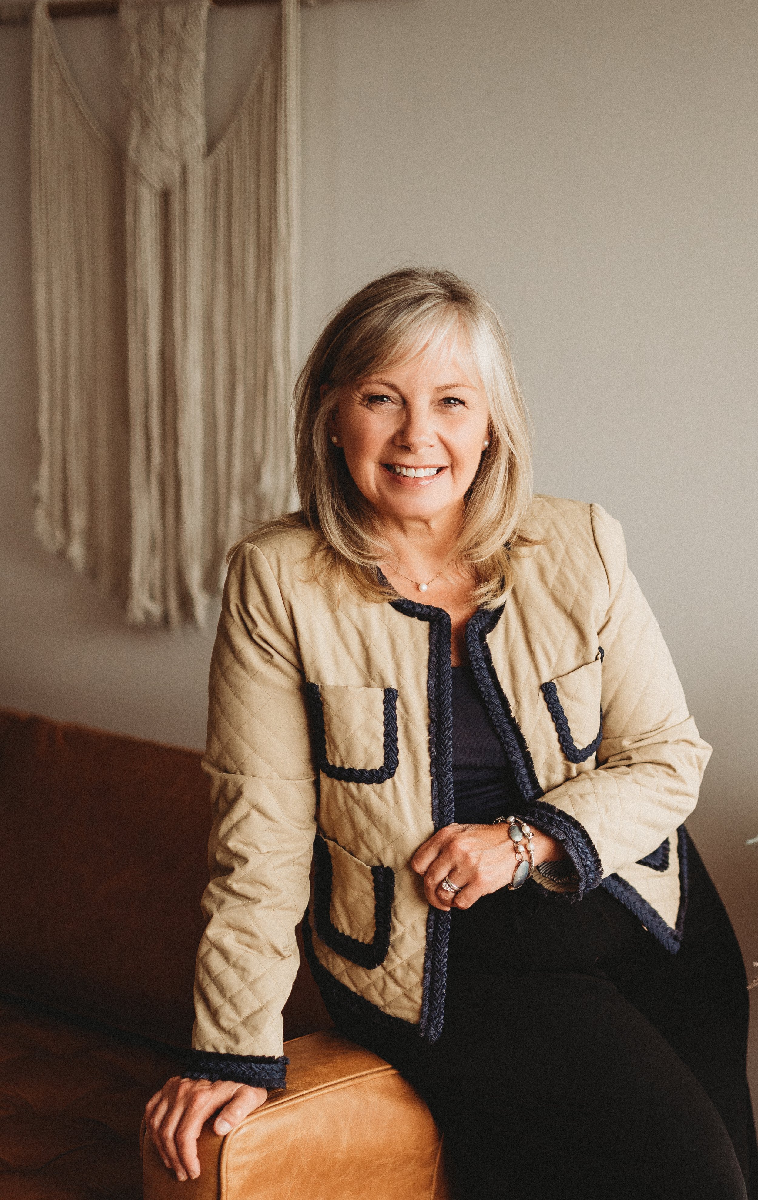 Woman sitting on a couch, wearing a beige quilted jacket with black trim, smiling, with macrame wall hanging in background.