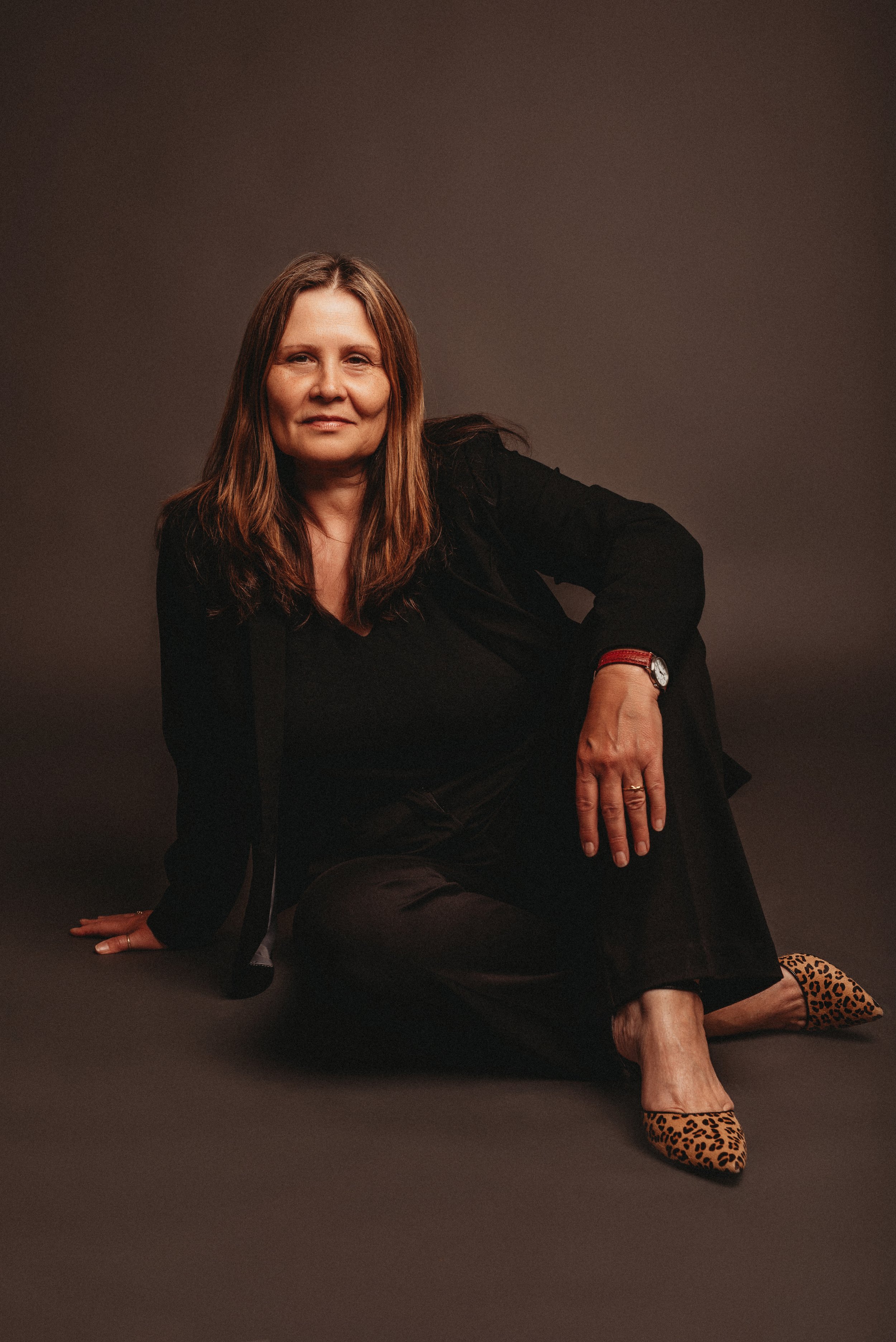 Person sitting on the floor wearing black outfit and leopard print shoes against a dark background.