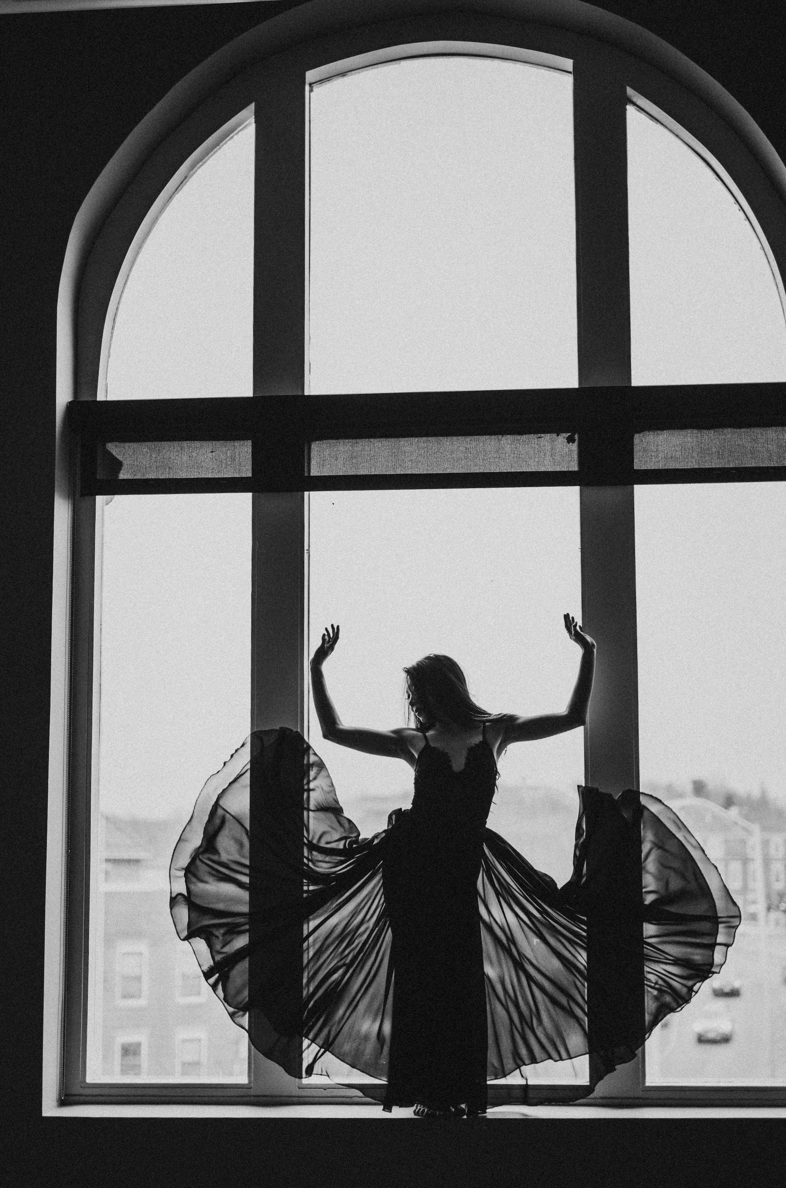 Silhouette of woman in flowing dress posing in front of large window.