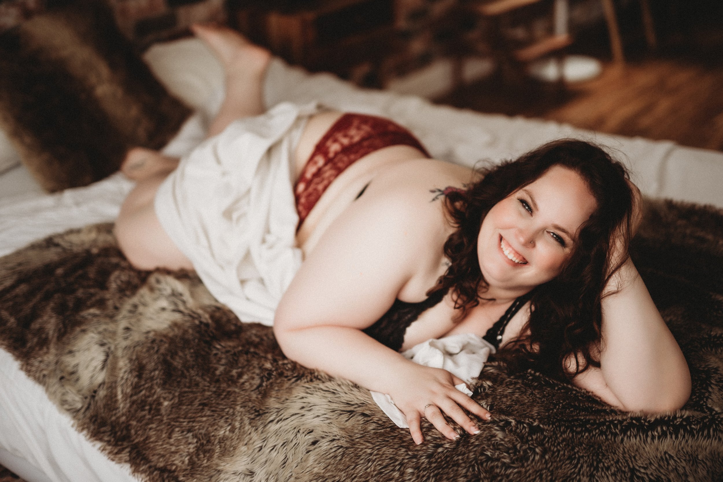 Smiling woman lying on bed with white sheet and fur blanket, wearing lace lingerie.