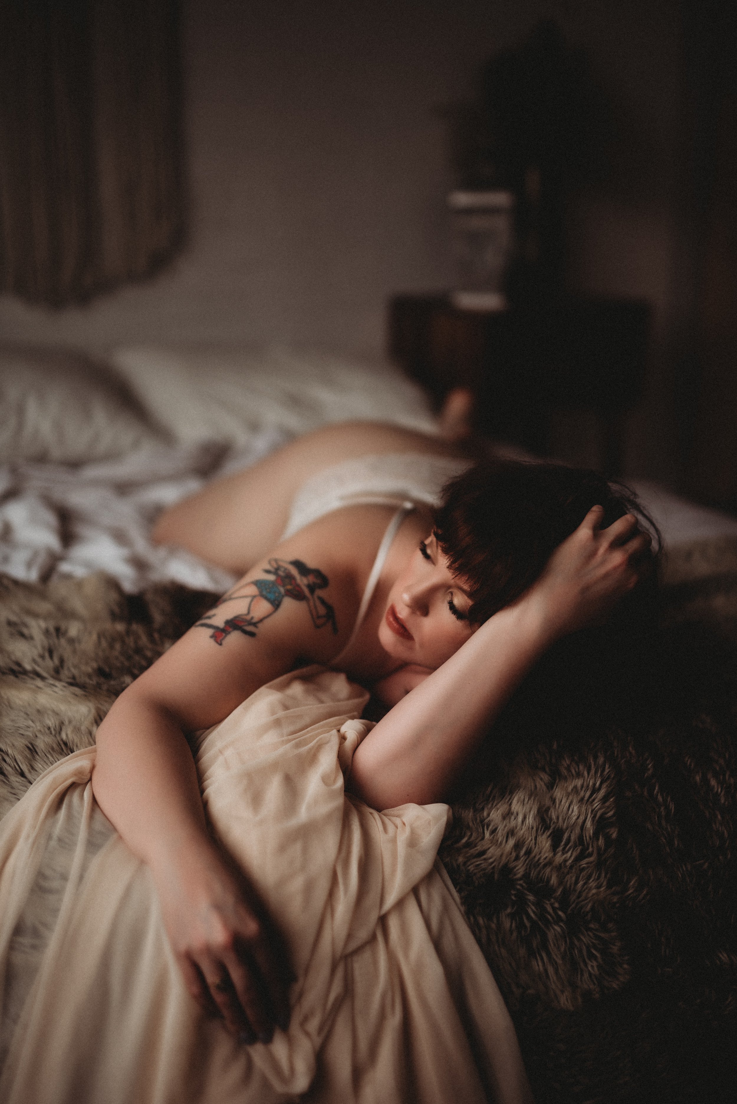 Woman with tattoo lying on bed, wearing white lingerie, wrapped in a beige sheet.