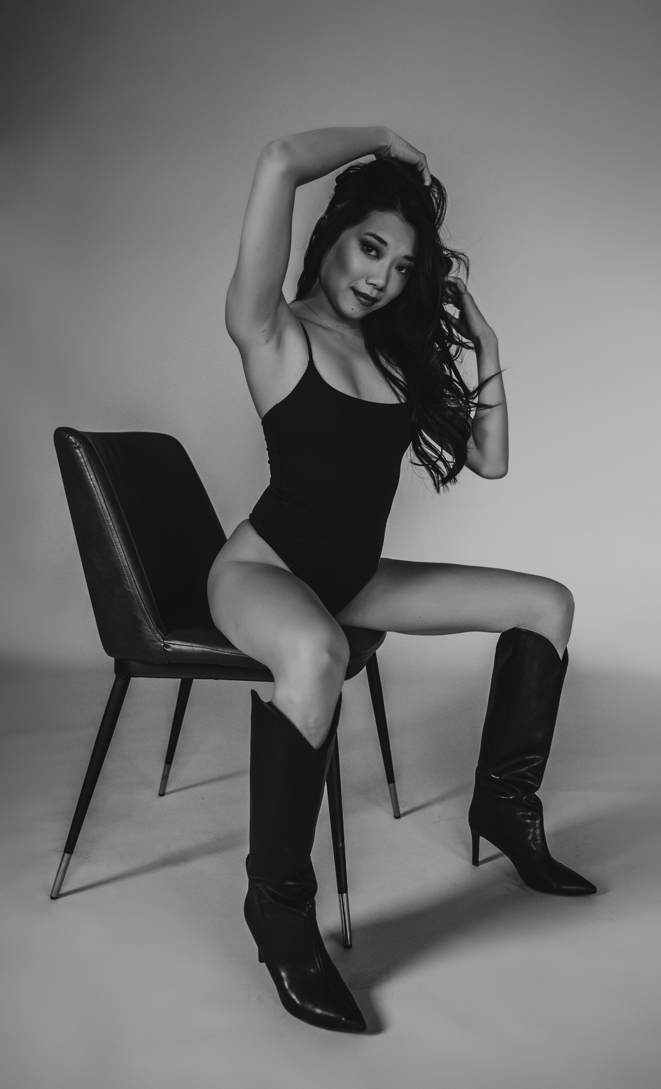 Black and white photo of a woman sitting on a chair, wearing a black bodysuit and knee-high boots, with one arm raised.