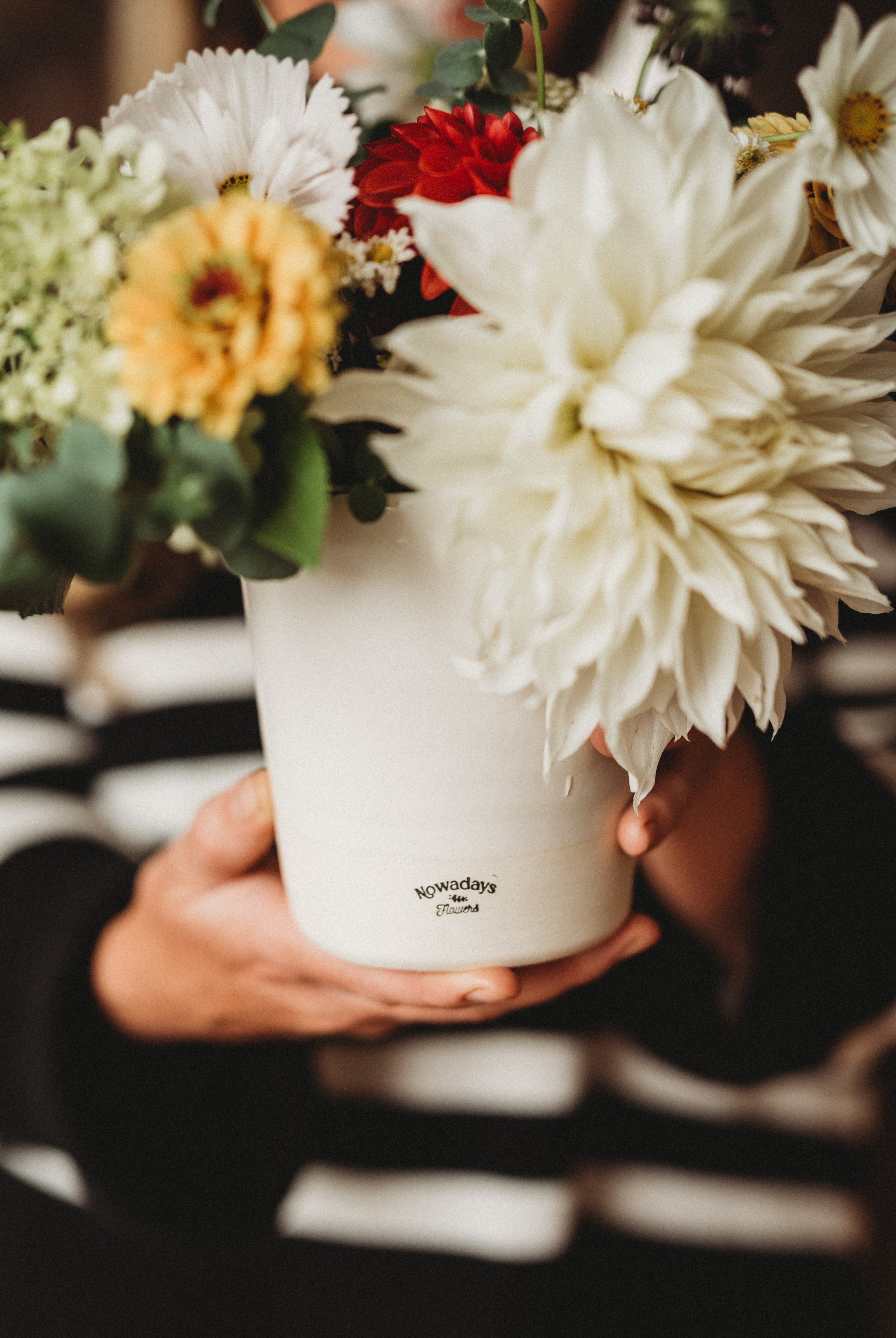A person holding a white ceramic pot with mixed flowers, including white, yellow, and red blooms, labeled 'Nowadays the Flower.'"