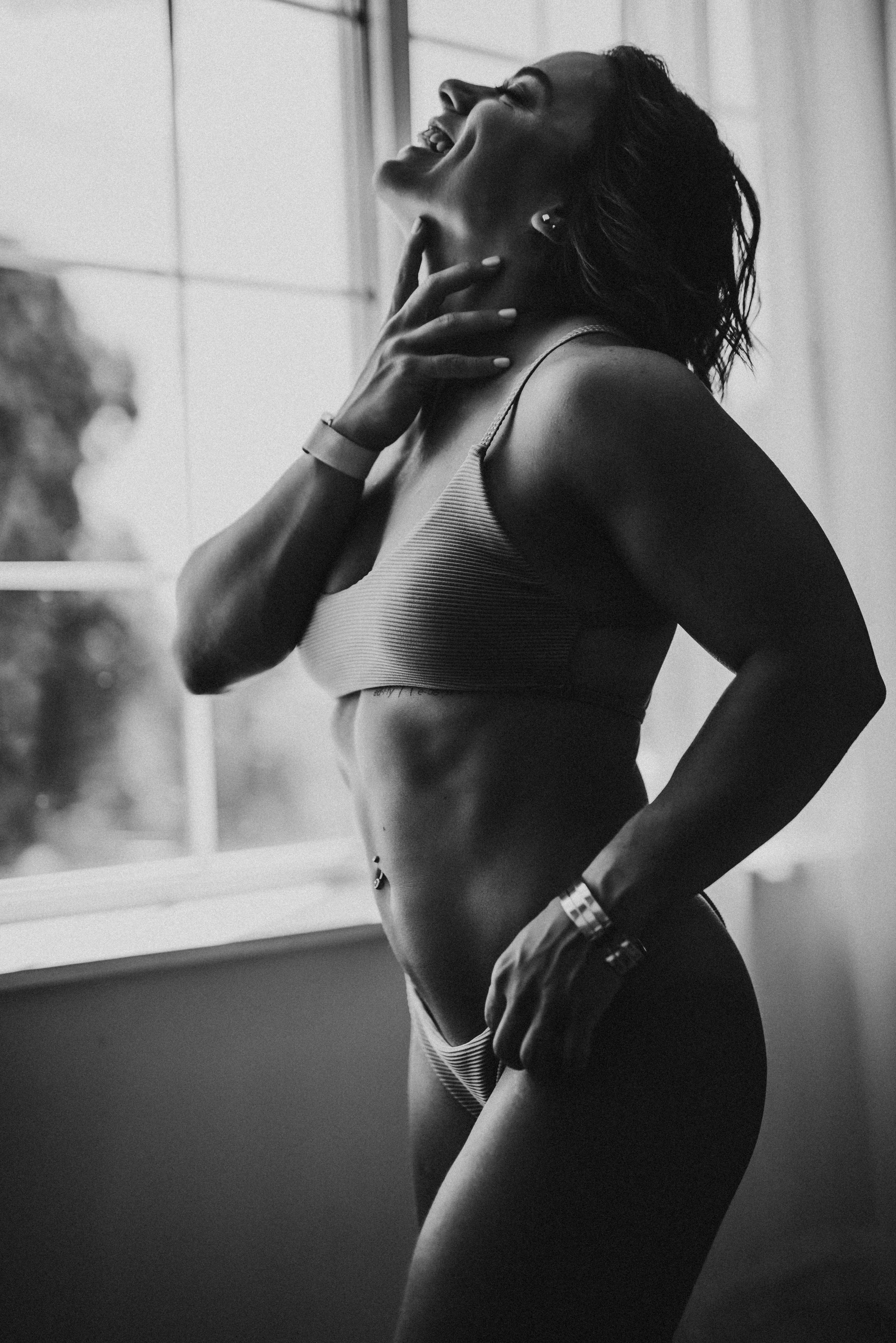 Black and white photo of a woman in underwear, smiling and touching her neck, standing by a window.