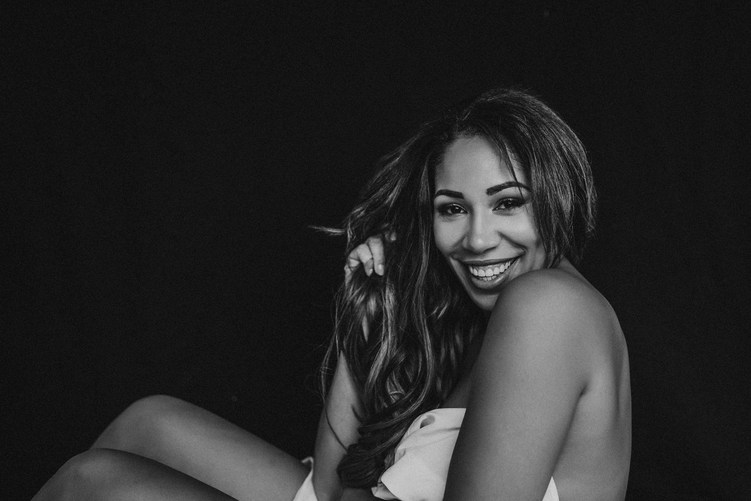 Black and white image of a smiling woman with long hair, sitting wrapped in a fabric against a dark background.