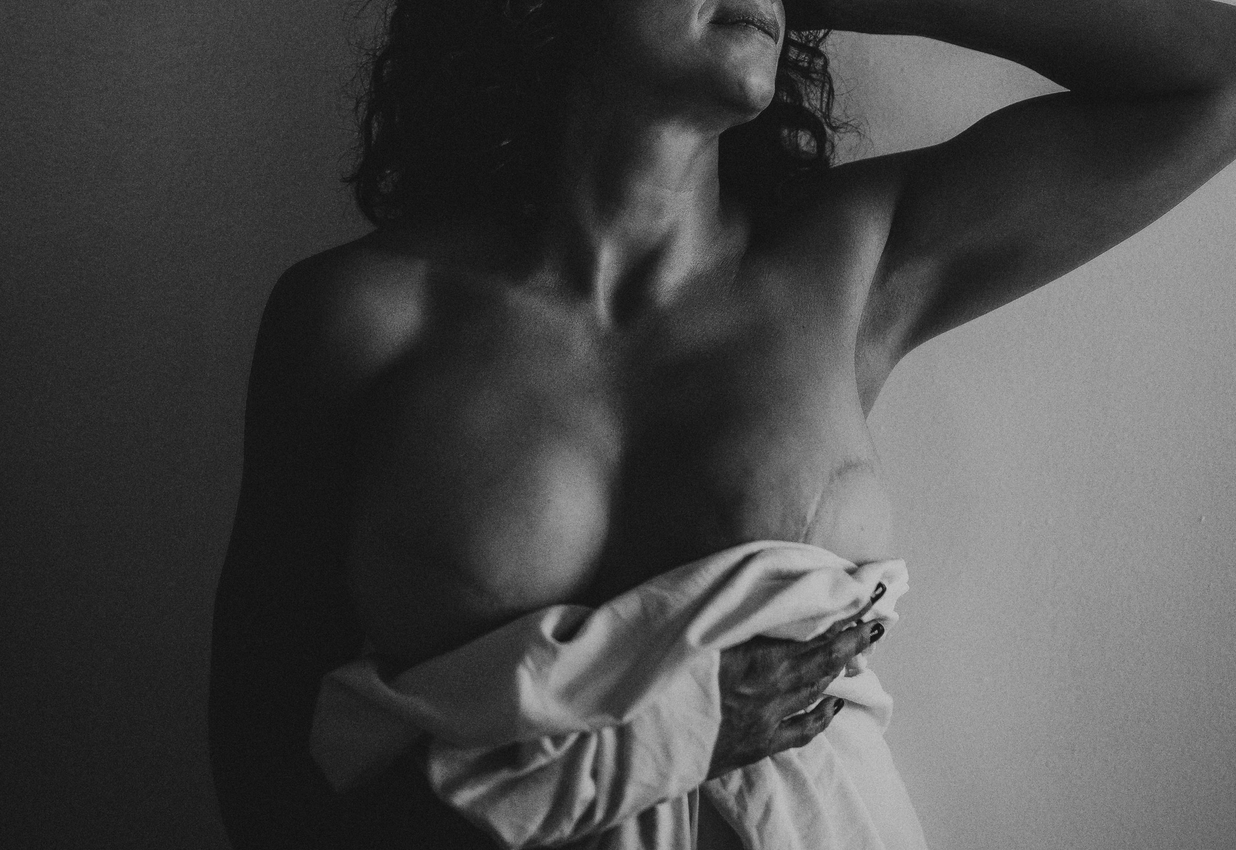 Black and white photograph of a partially covered person, focusing on the torso and hand holding a draped cloth. The image emphasizes shadows and contours.