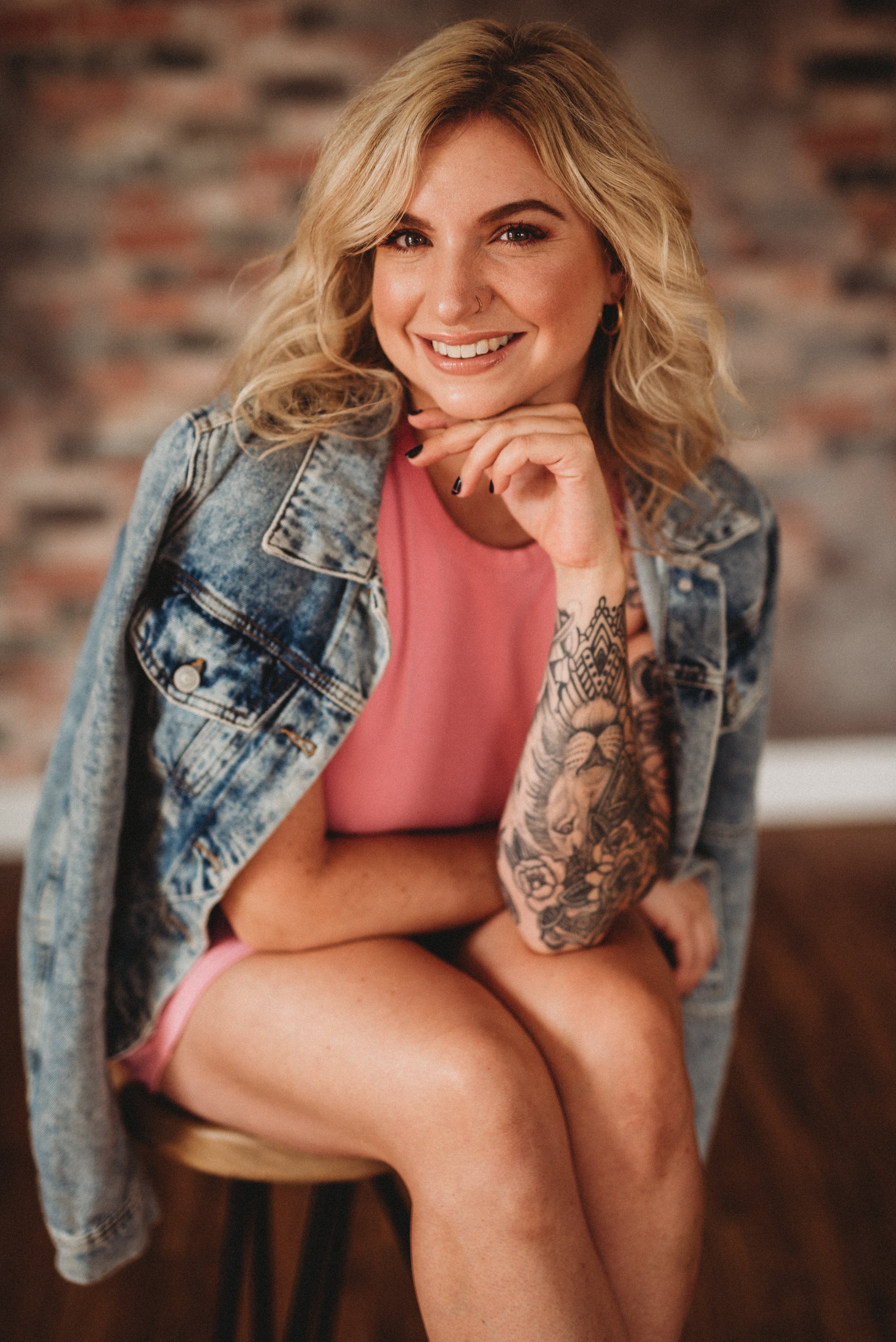 Woman with blonde hair smiling, wearing pink dress and denim jacket, with tattooed arm.