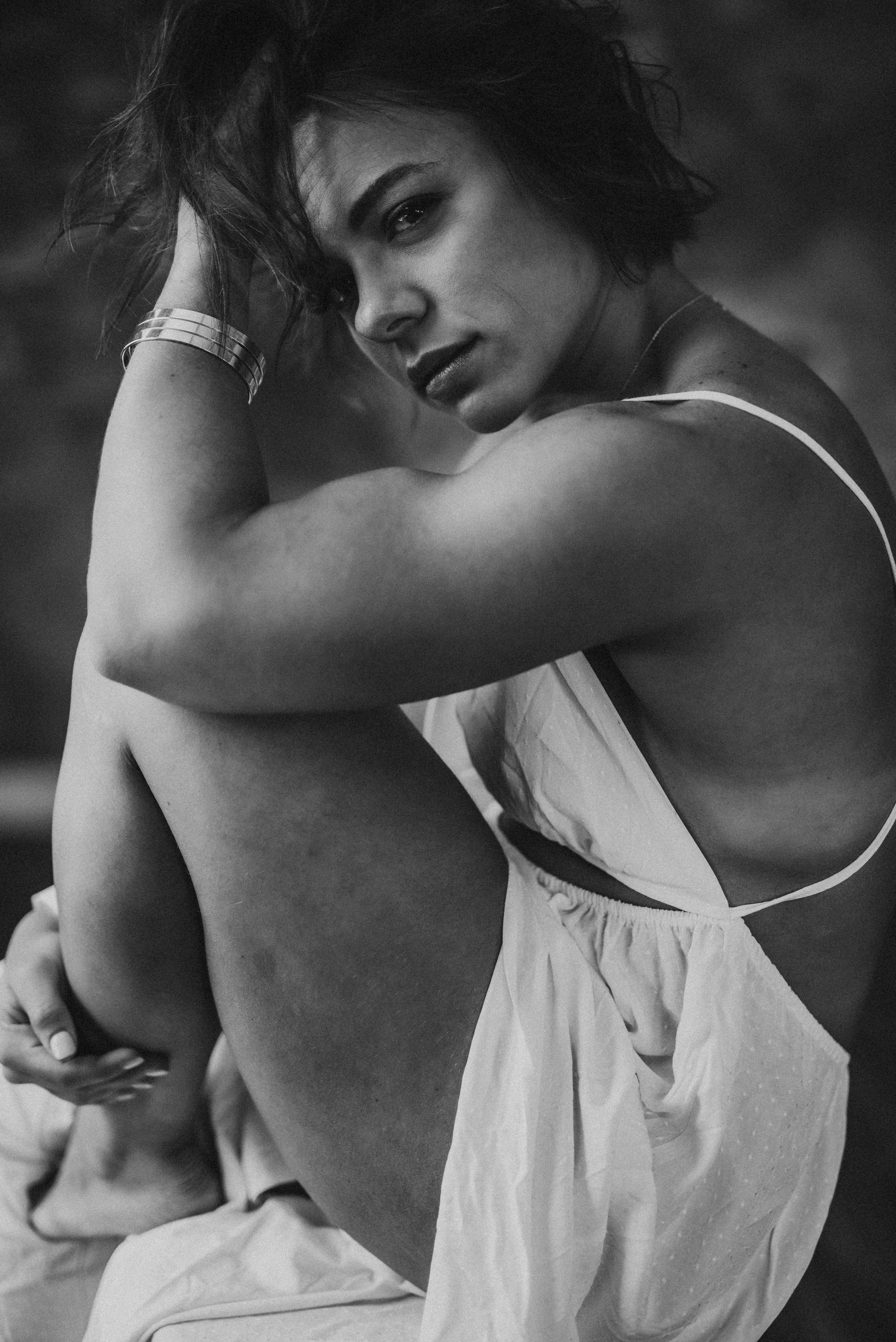 Black and white portrait of a woman sitting, wearing a sleeveless dress, with her legs drawn up and hand in her hair.