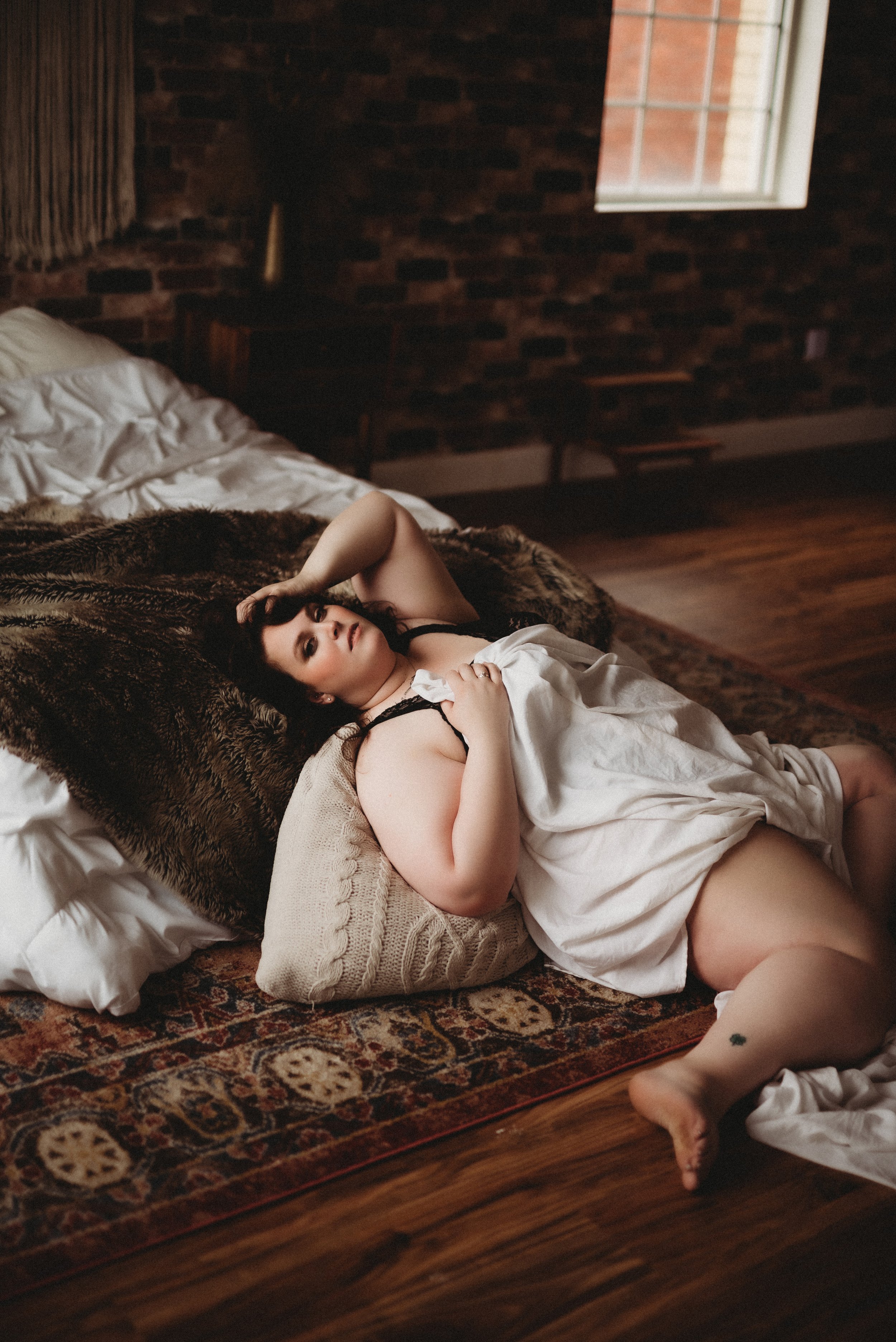 Person lounging on a rug with pillows, draped in a white sheet, in a room with a brick wall and window.
