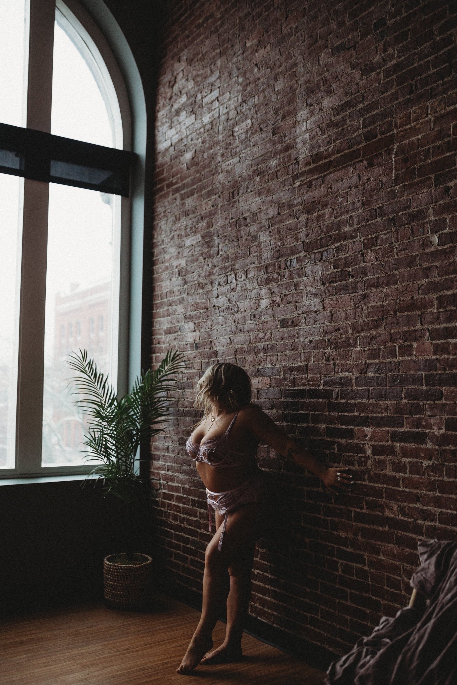 Person in lingerie standing against a brick wall with large window and potted plant nearby.
