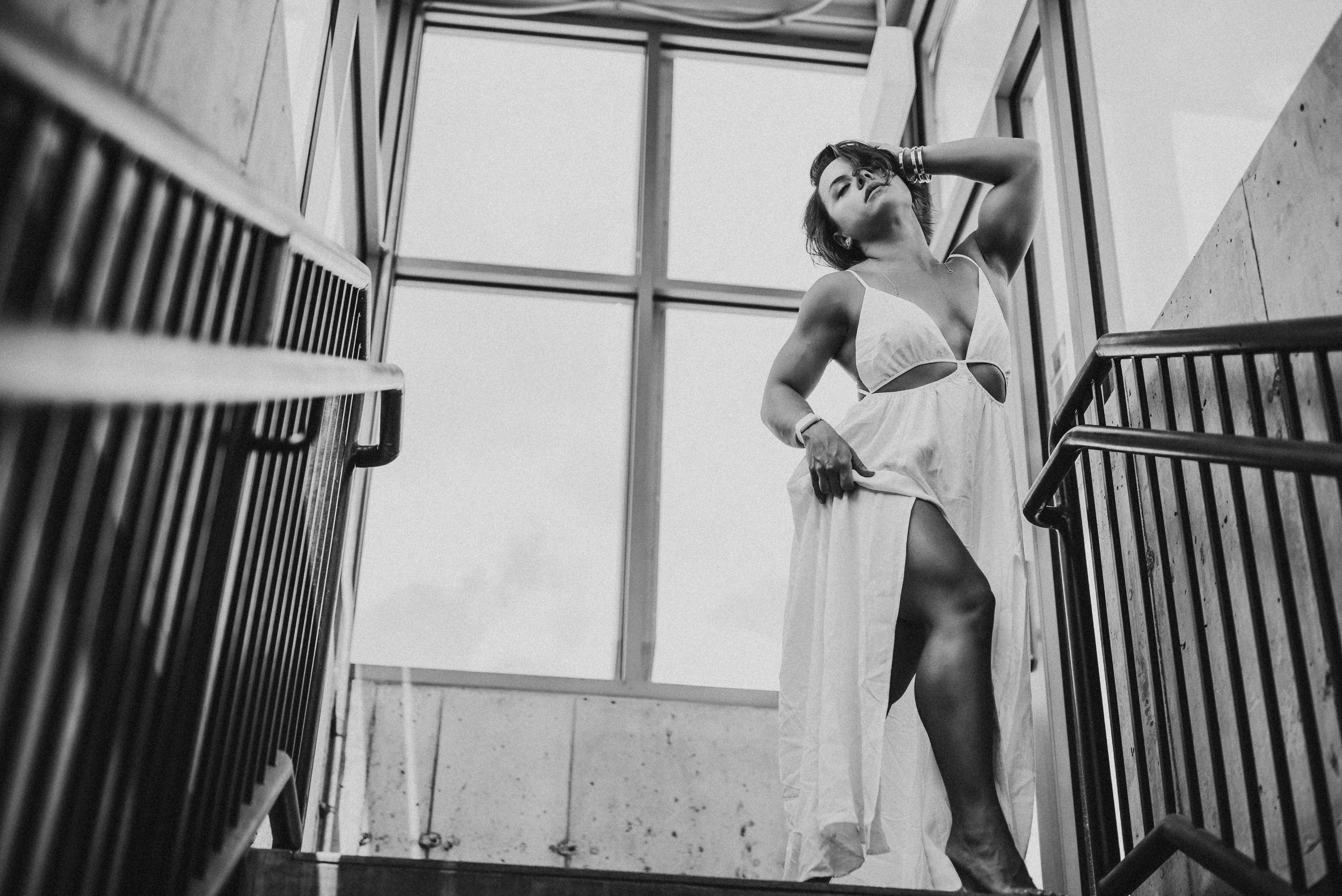 Black and white photo of a woman in a white dress posing on stairs with windows in the background.