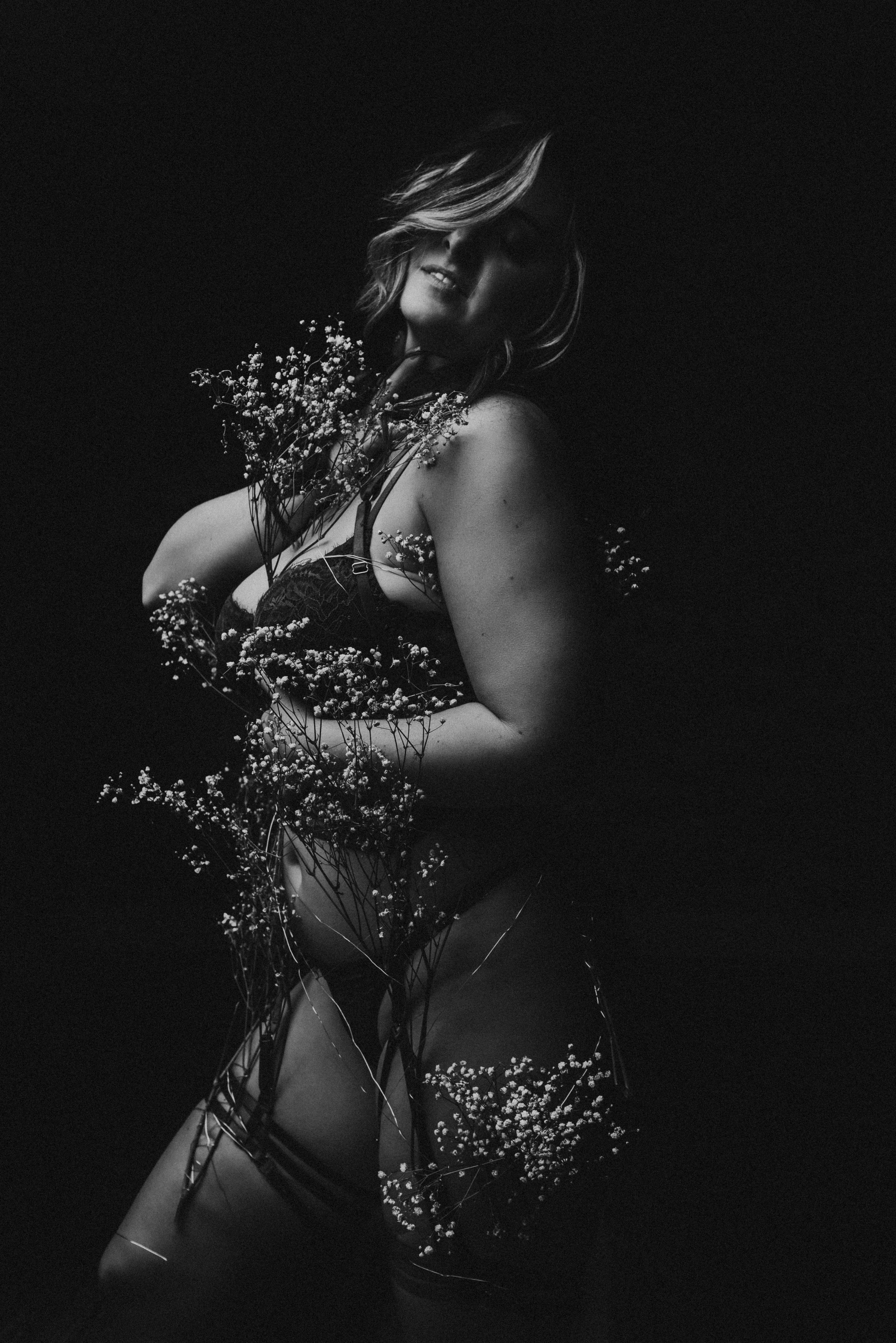 Black and white photo of a person in lingerie adorned with flowers against a dark background.