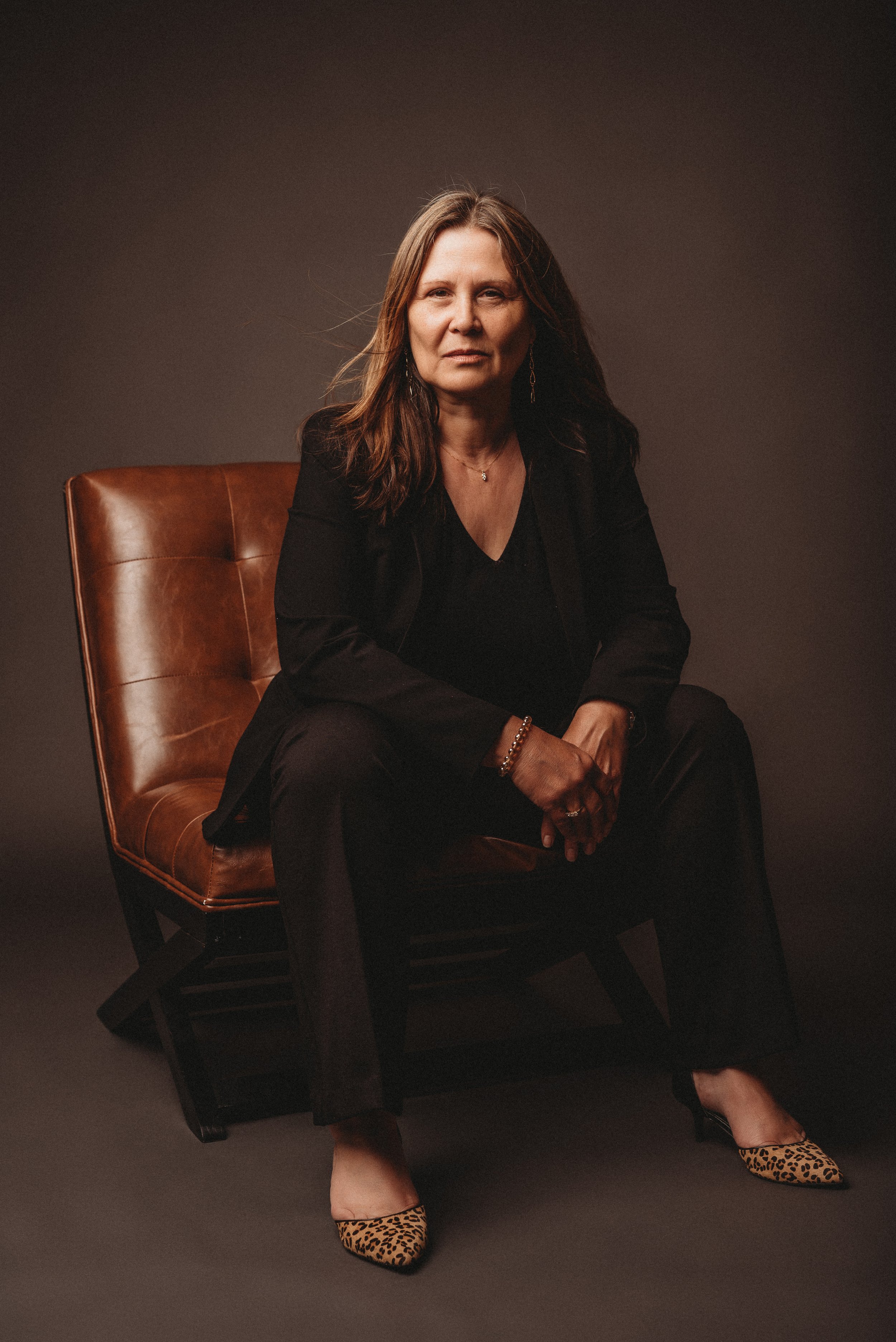 Person sitting on a leather chair in a dark room, wearing a black suit and leopard print shoes.