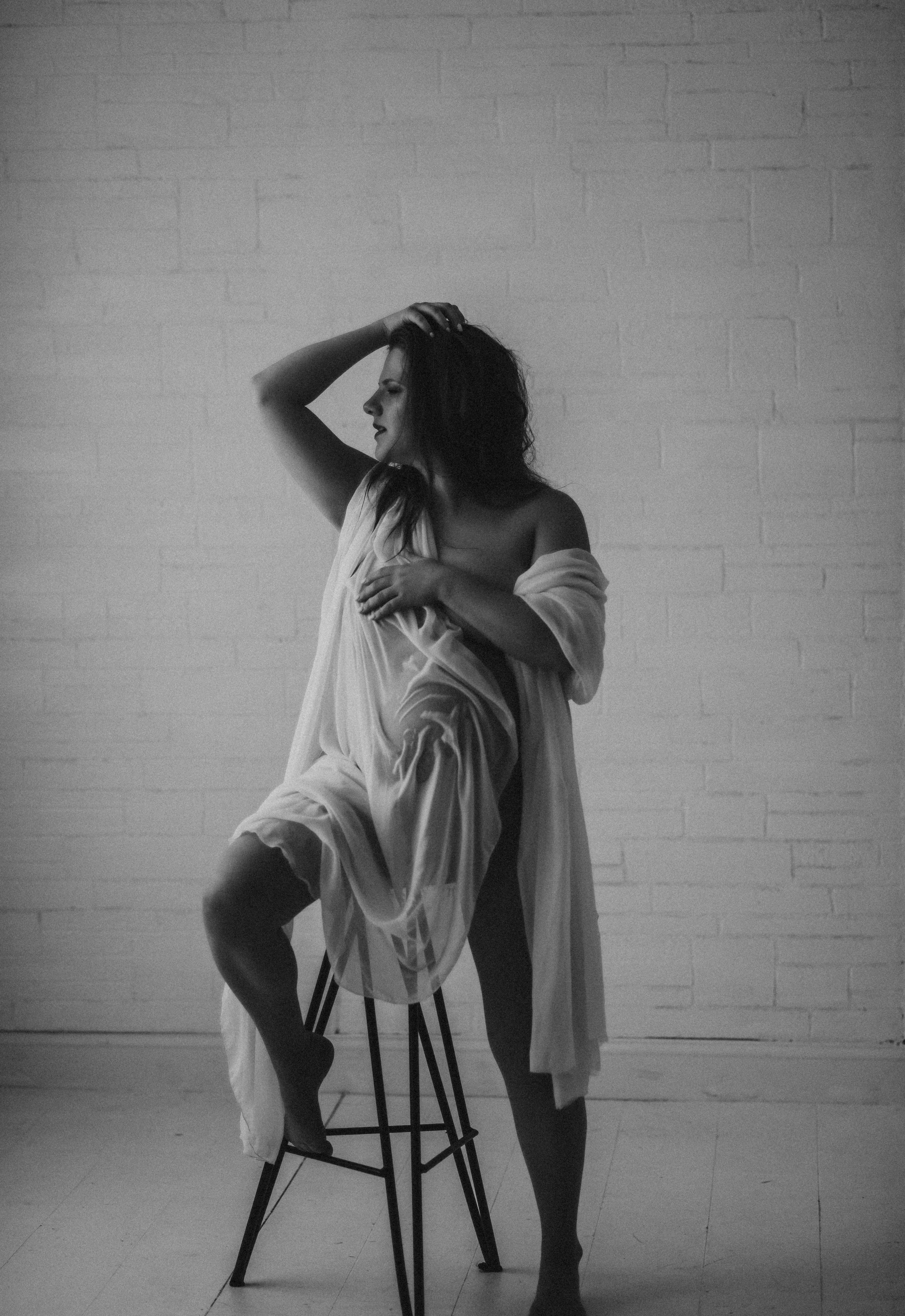 Black and white photo of a woman with long hair sitting on a stool, draped in a sheer fabric, posing against a brick wall.