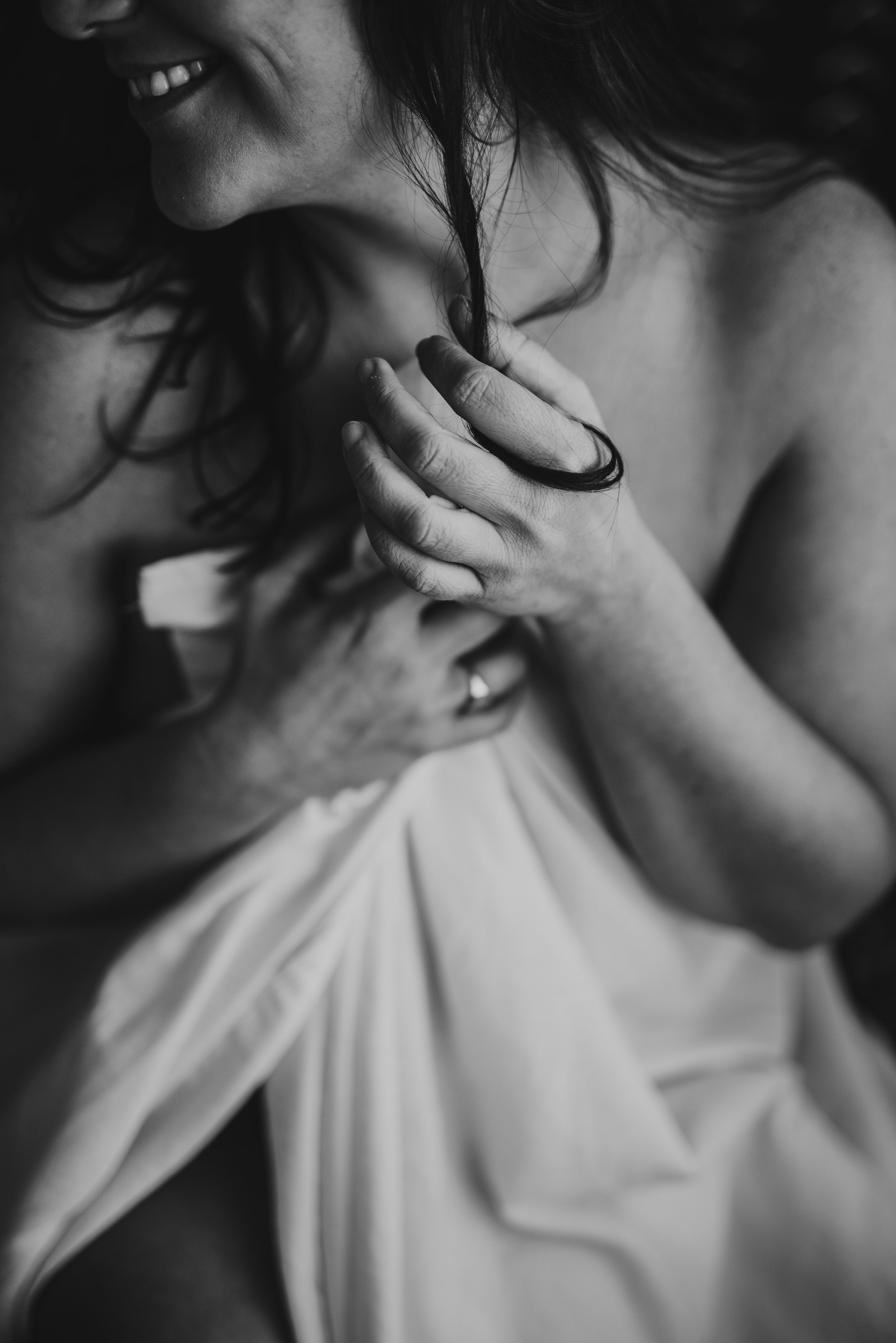 Black and white close-up of a woman smiling, holding a strand of hair, partially covered with a sheet.