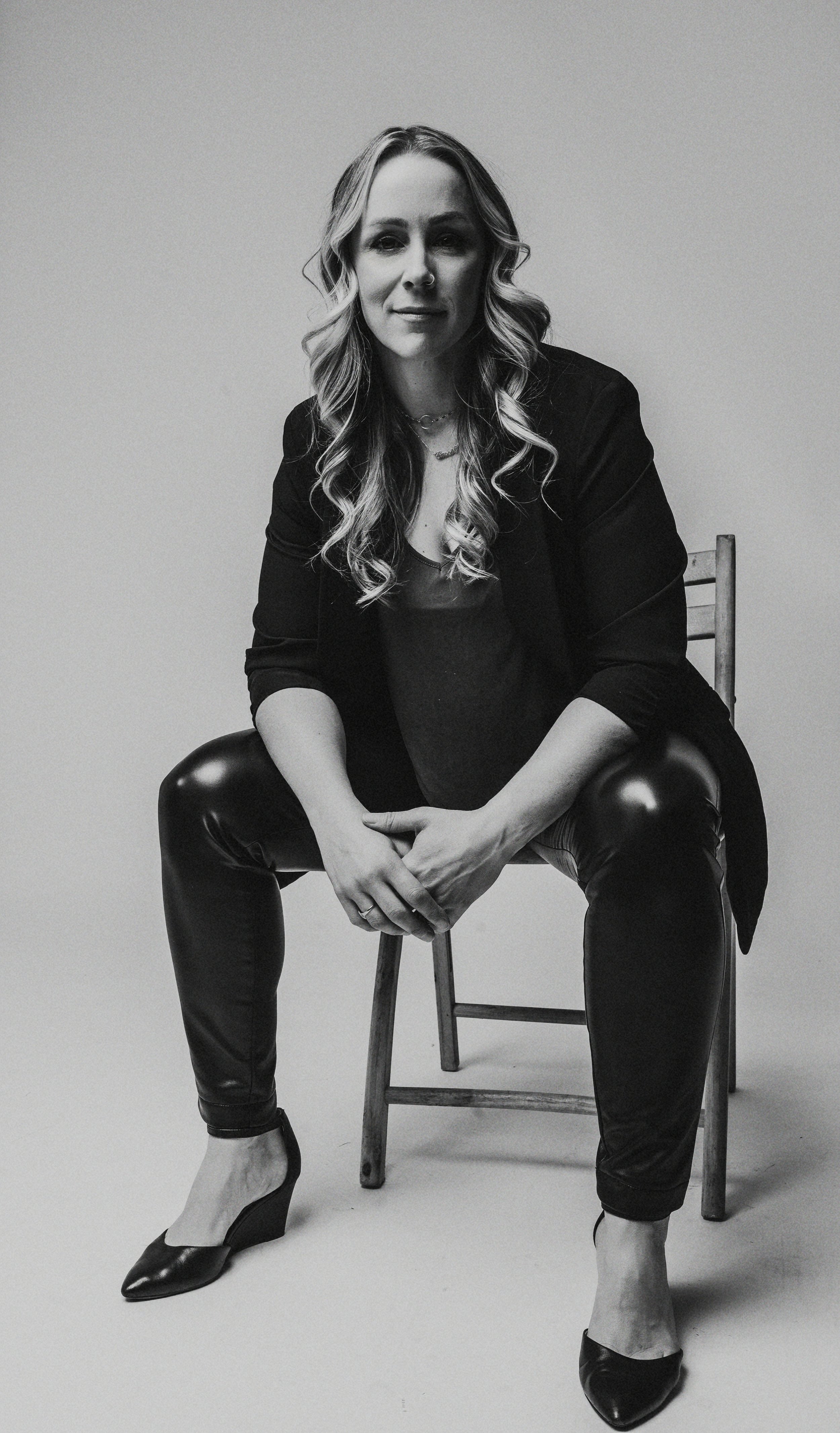 Black and white photo of a woman seated on a chair in leather pants and a blazer, with long wavy hair, looking at the camera.
