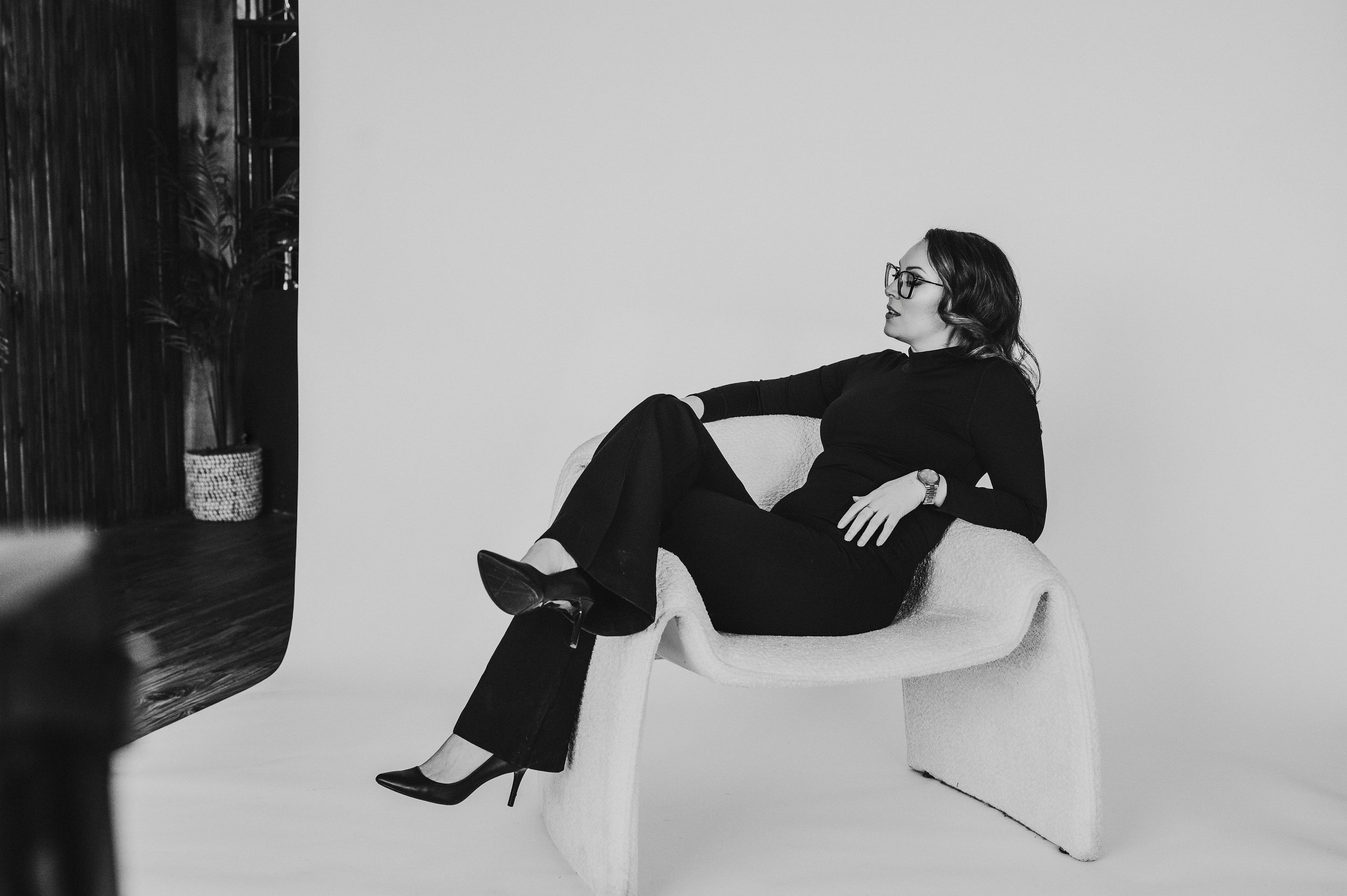 Black and white photo of a woman sitting in a modern chair, wearing dark clothing and glasses, legs crossed. Indoor setting with a potted plant in the background.
