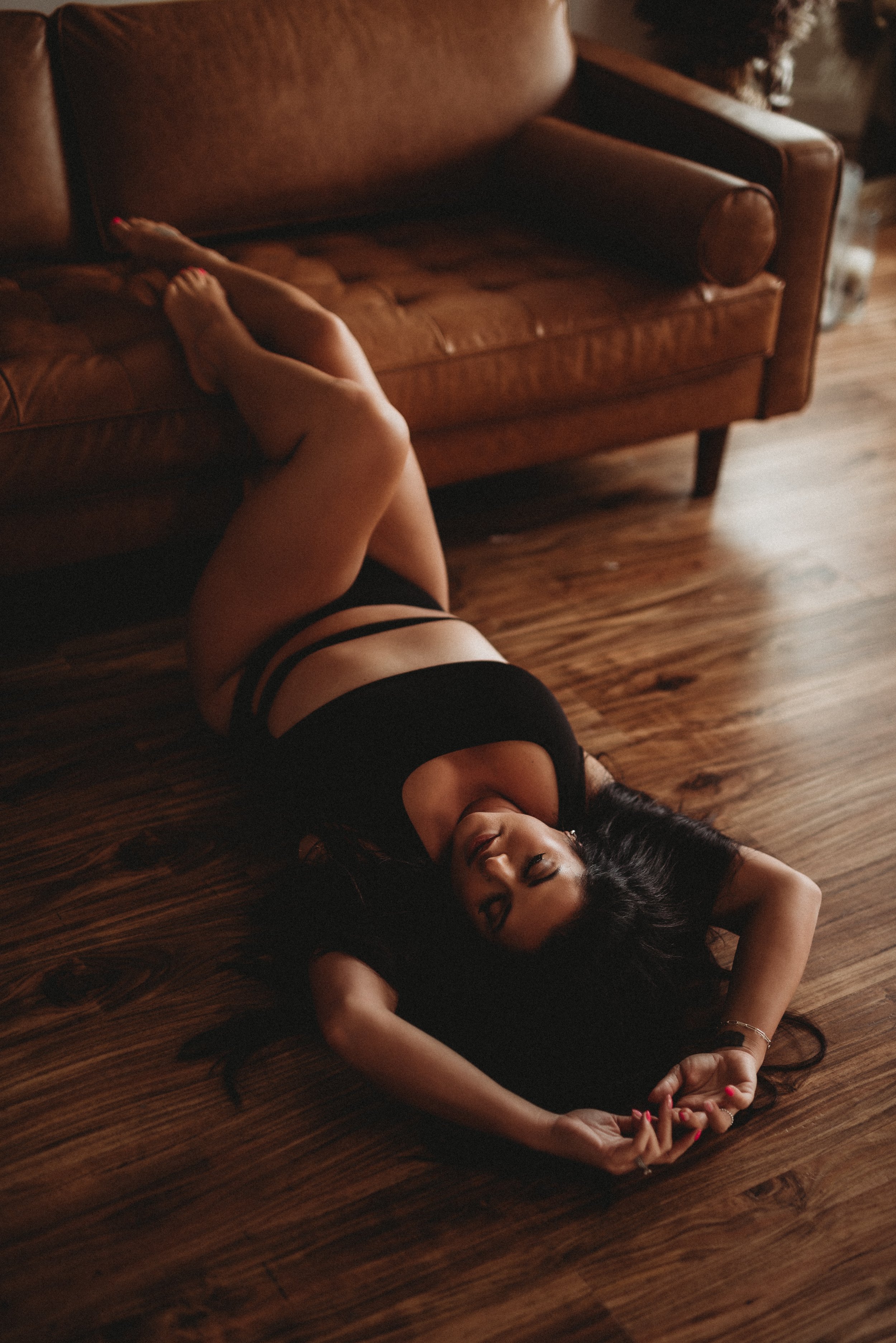 Person in black outfit lying on wooden floor with head near a brown leather sofa, arms extended above head.