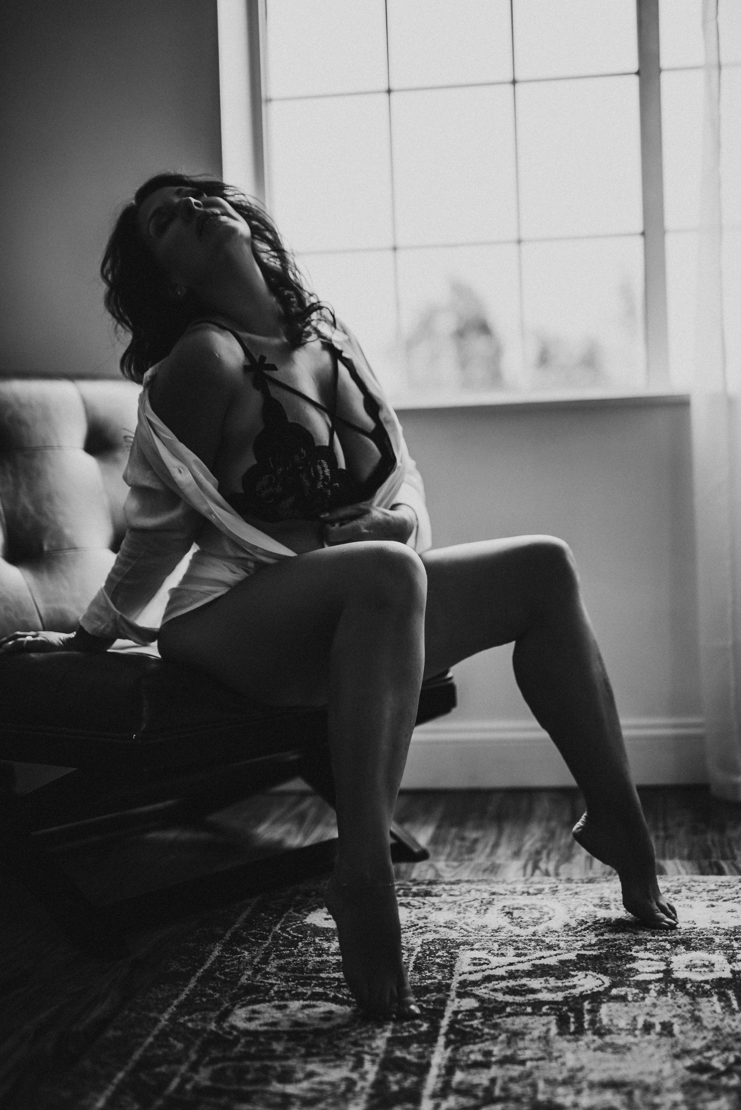 Black and white photo of a woman sitting on a chair wearing lingerie, with natural light coming through a window.