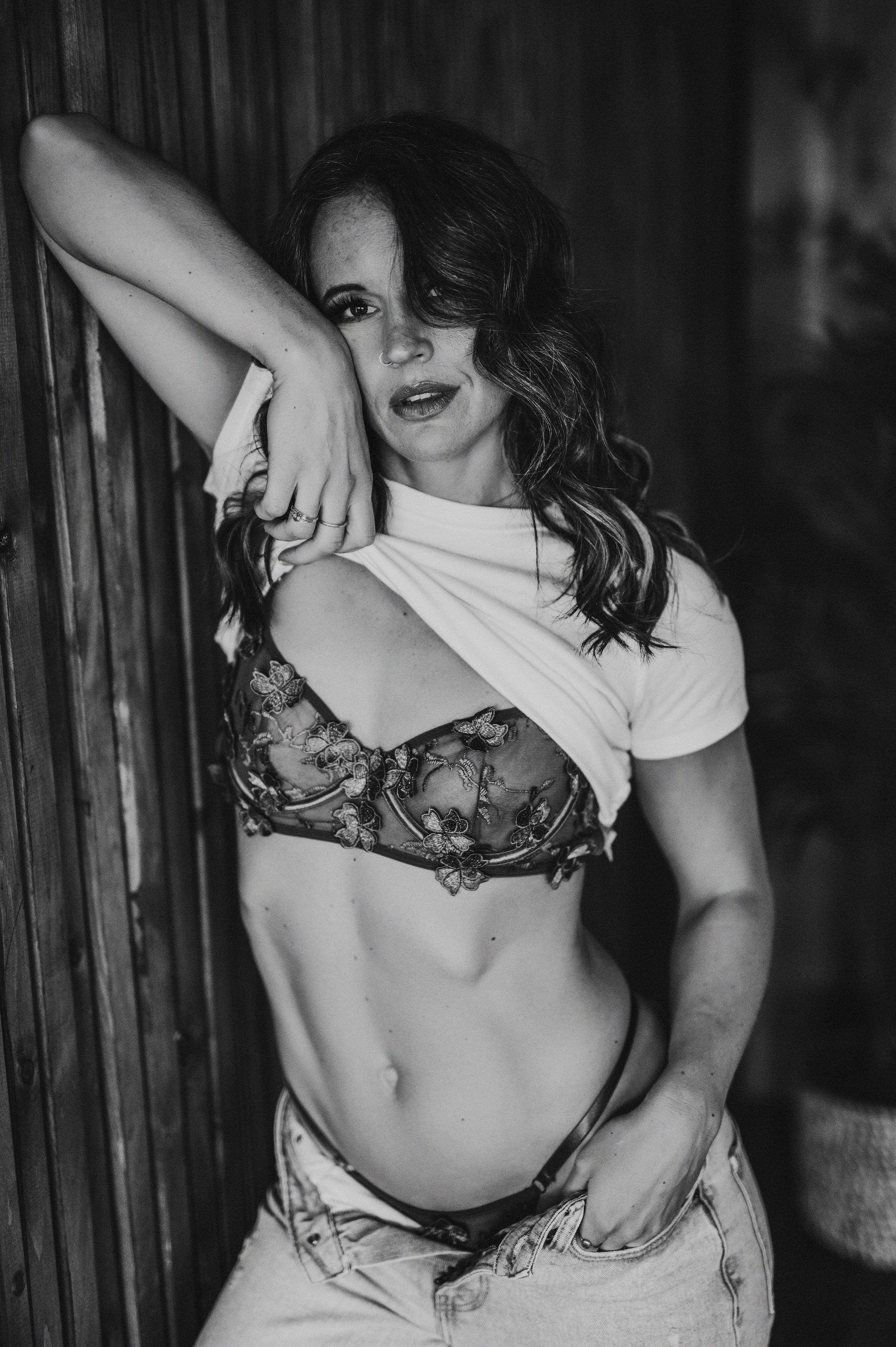 Black and white portrait of a woman in a floral bra and jeans, lifting her shirt, in front of a wooden wall.
