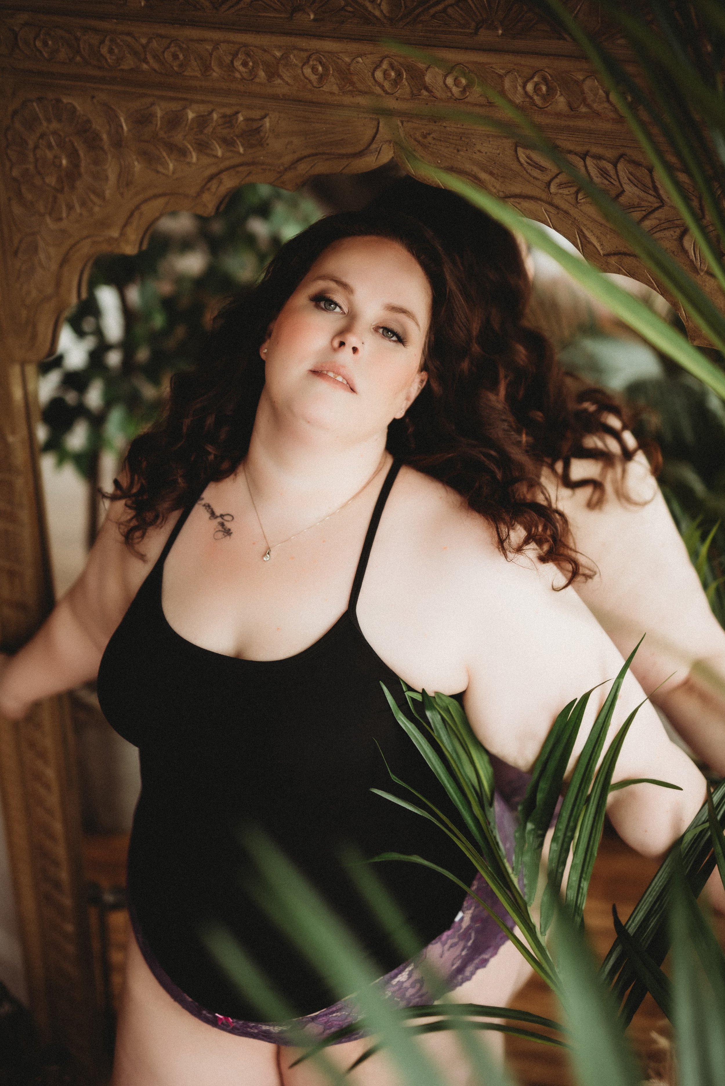 Person in a black tank top and purple lace underwear posing in front of an ornate mirror with green plant leaves in the foreground.