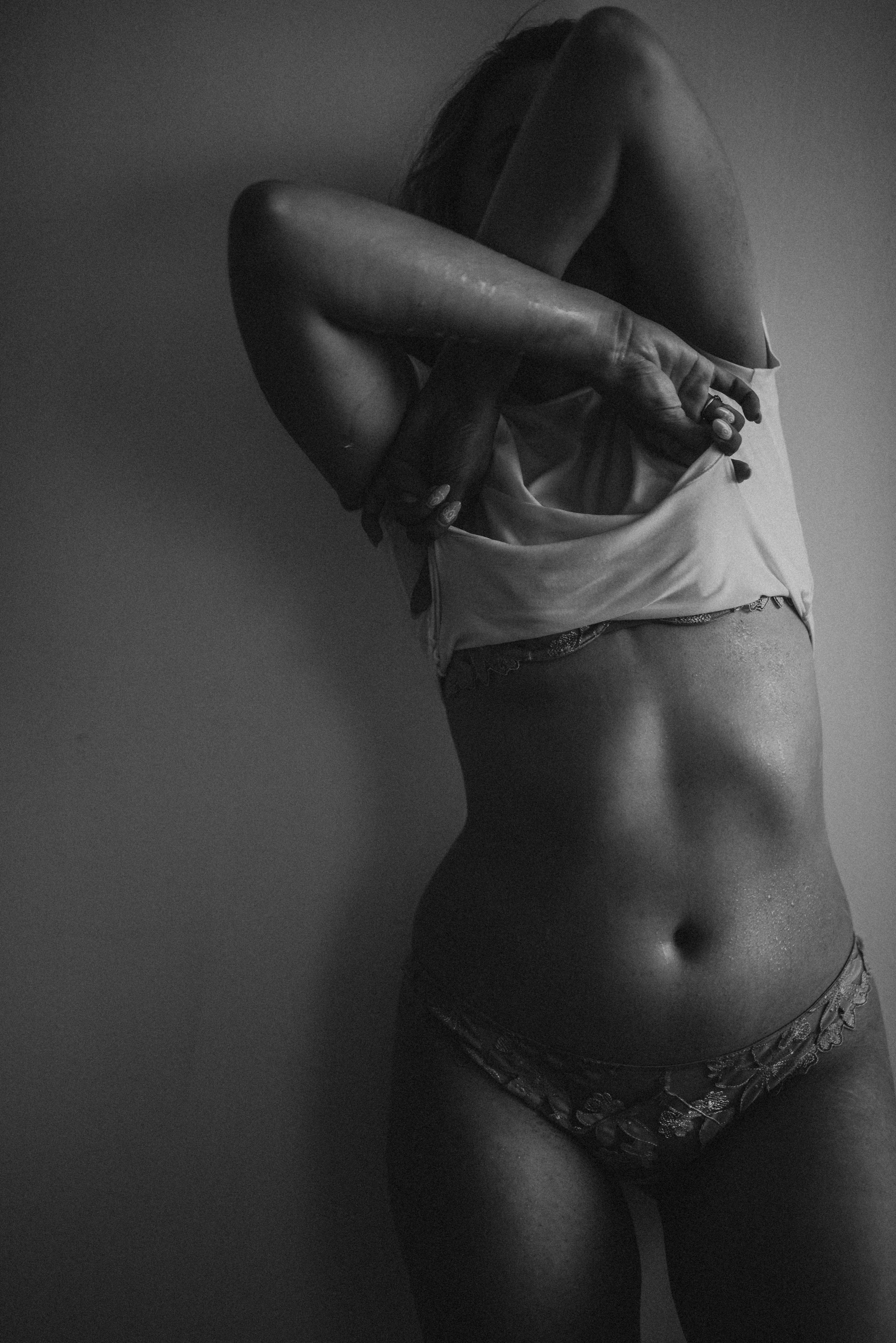 Black and white photo of a person standing against a wall, partially obscured as they lift their top, revealing their midriff and wearing patterned underwear.