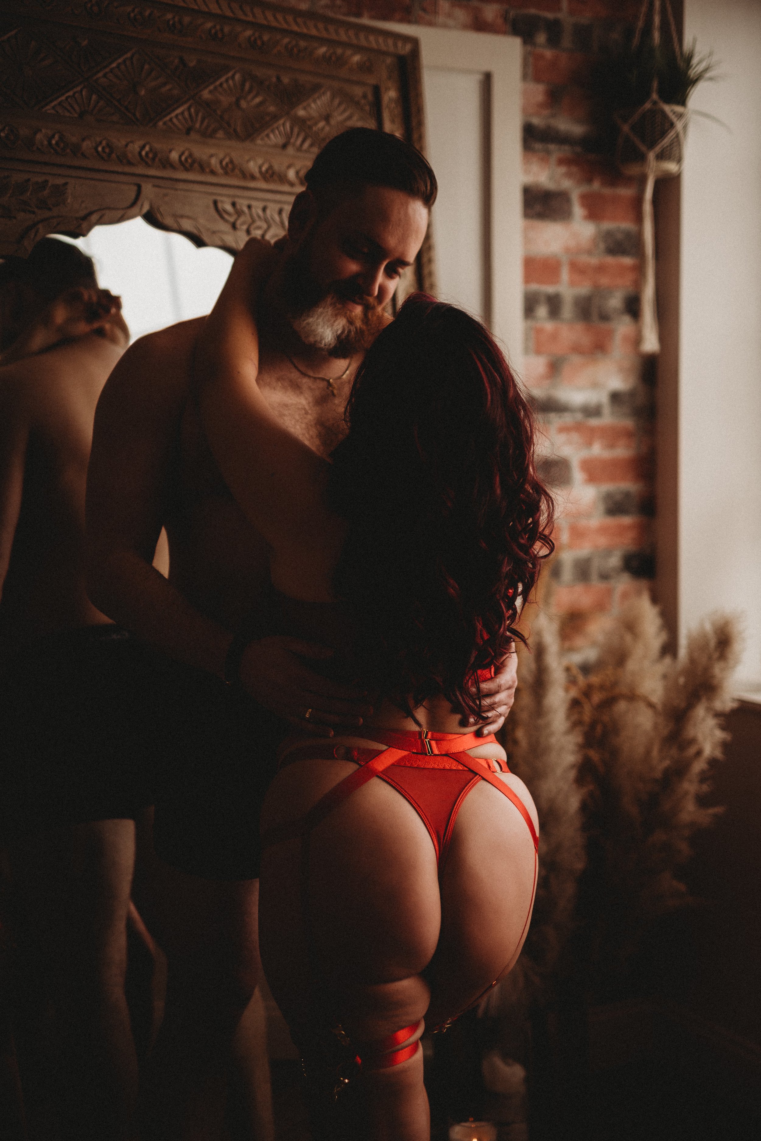 A couple embracing intimately in a dim room, with a decorative mirror and pampas grass in the background.
